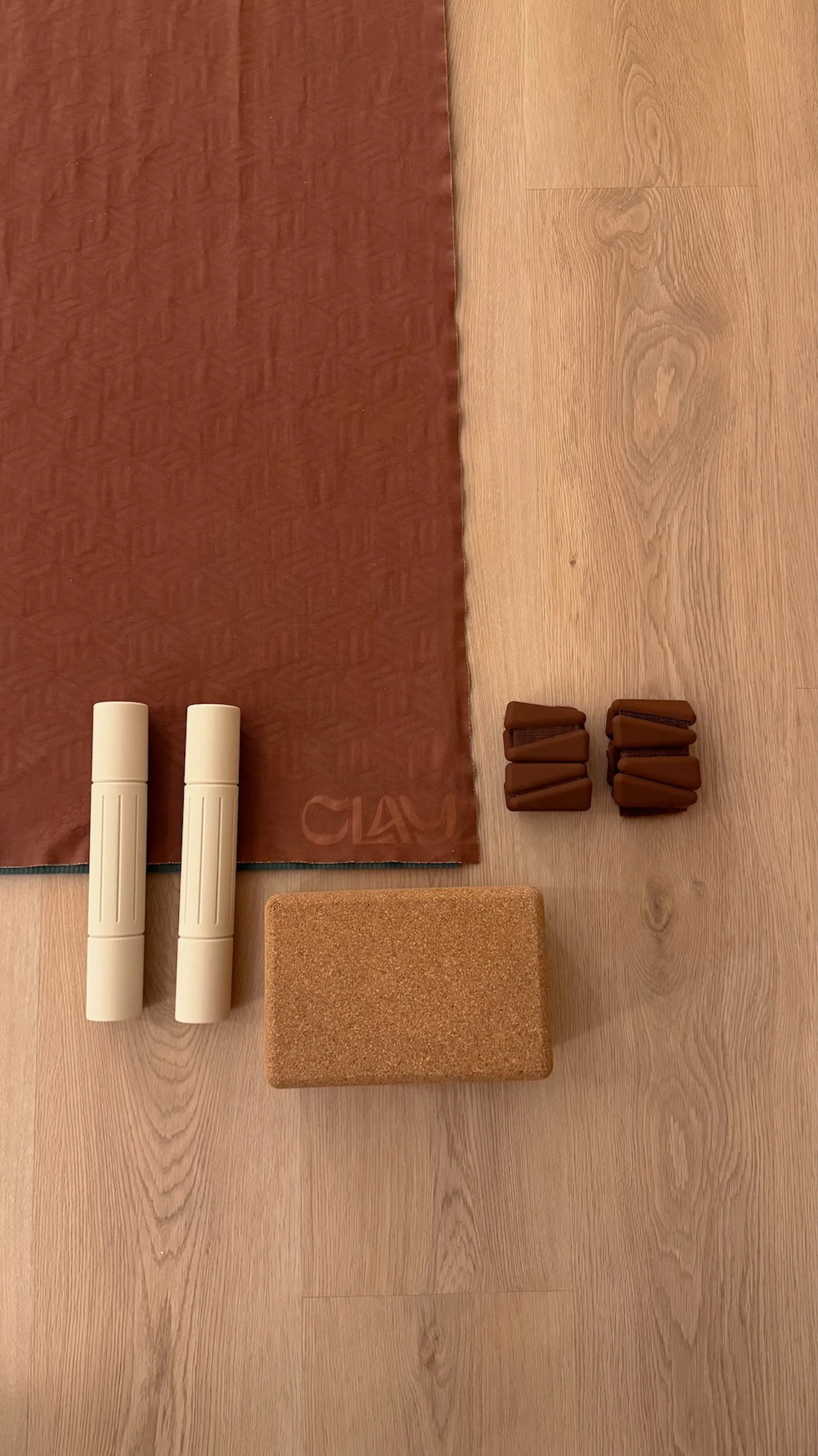 Brown textured yoga mat with "CLAY" logo, two beige foam grips, a cork yoga block, and two pairs of brown resistance bands on a light wooden floor.