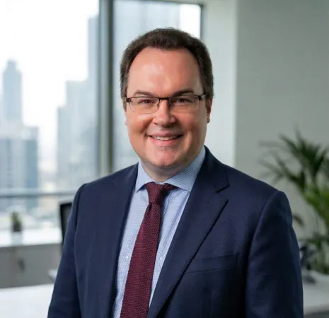 Man wearing glasses, a dark suit, blue shirt, and red tie, smiling in an office setting with a cityscape visible through the window behind him.