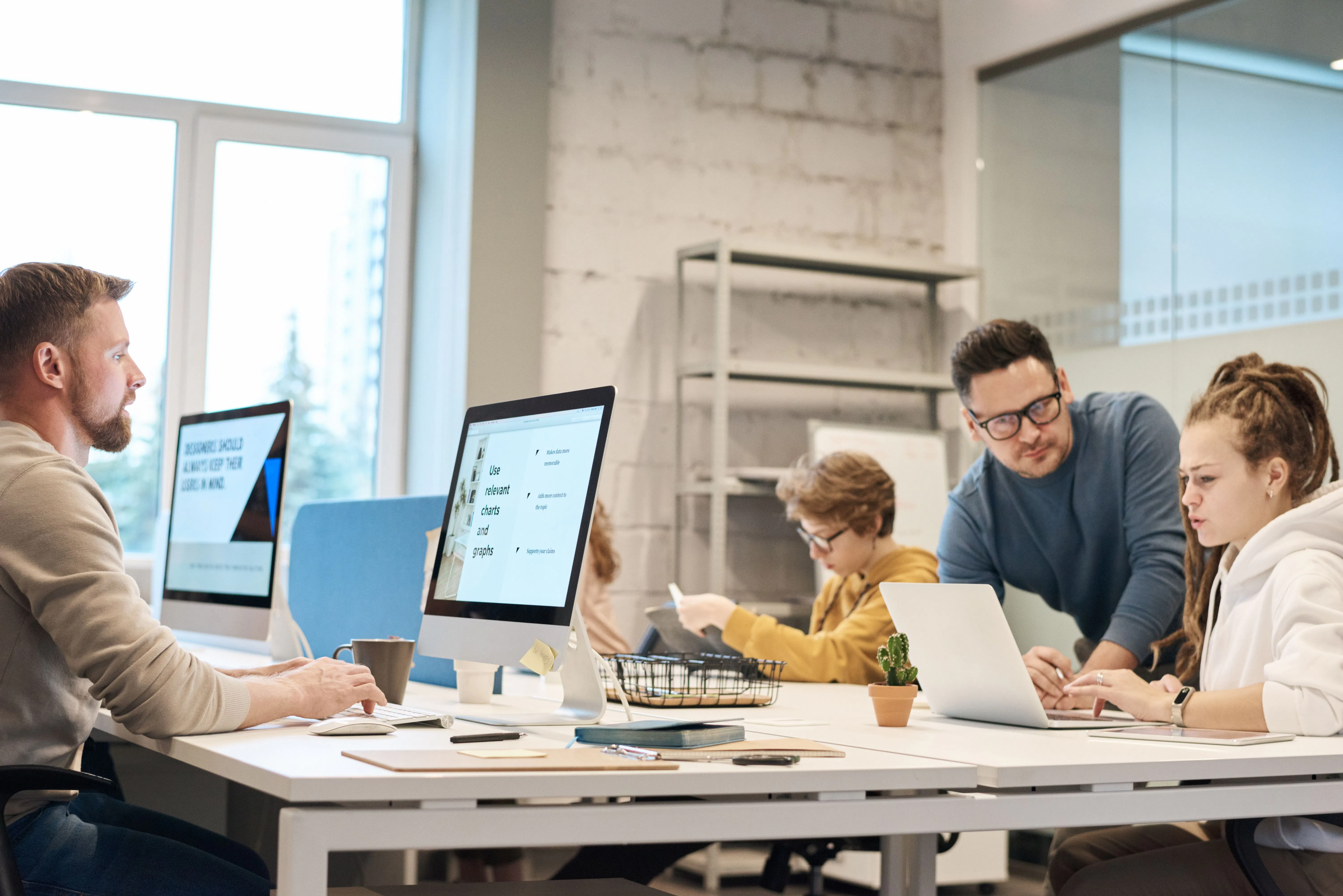 Vier Personen arbeiten in einem modernen Büro an Computern und einem Laptop, zwei diskutieren konzentriert, zwei arbeiten einzeln, helle Tageslichtumgebung.