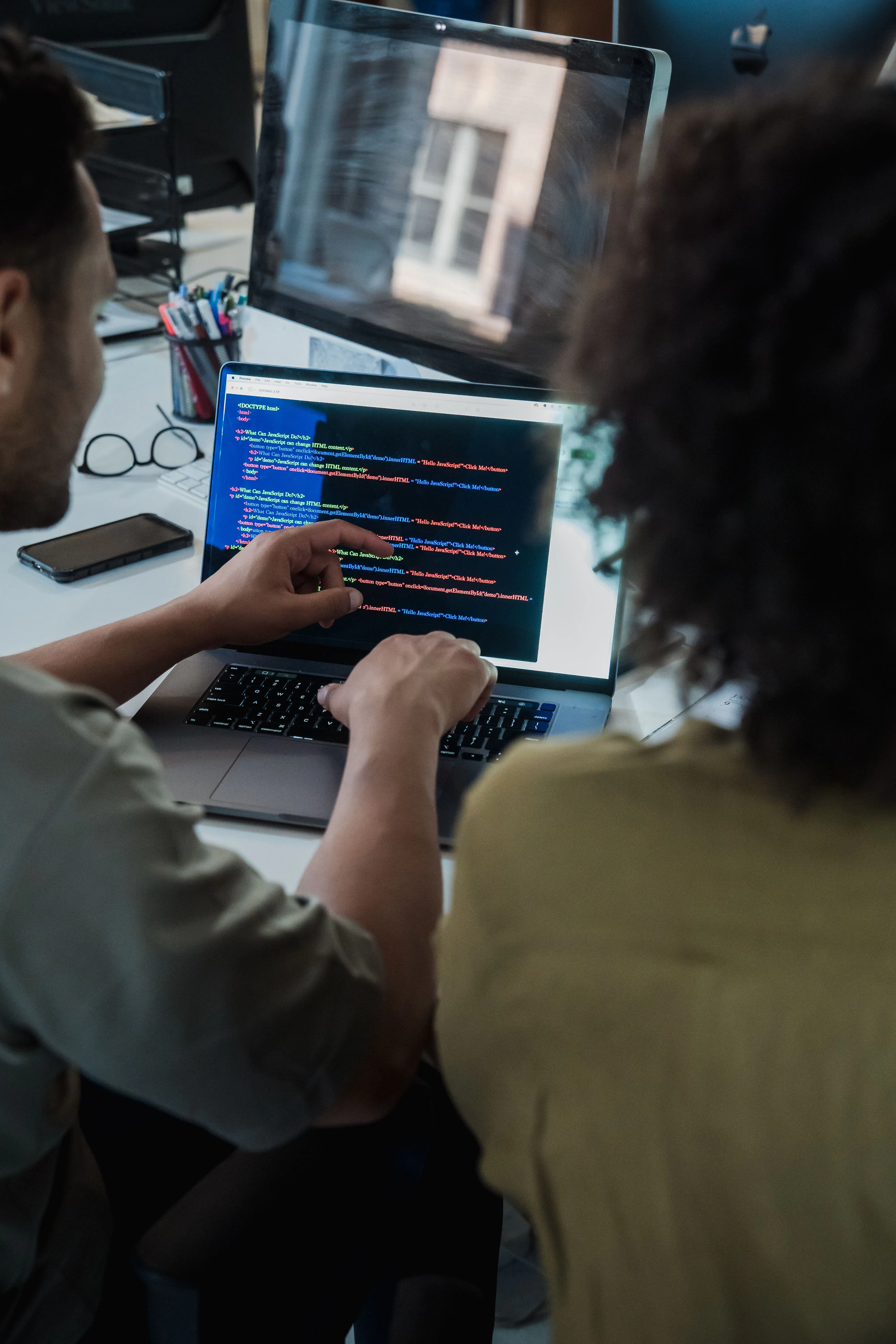 Zwei Personen betrachten und diskutieren Computerprogrammierung auf einem Laptopbildschirm in einem Büro mit Schreibutensilien und zwei Monitoren.