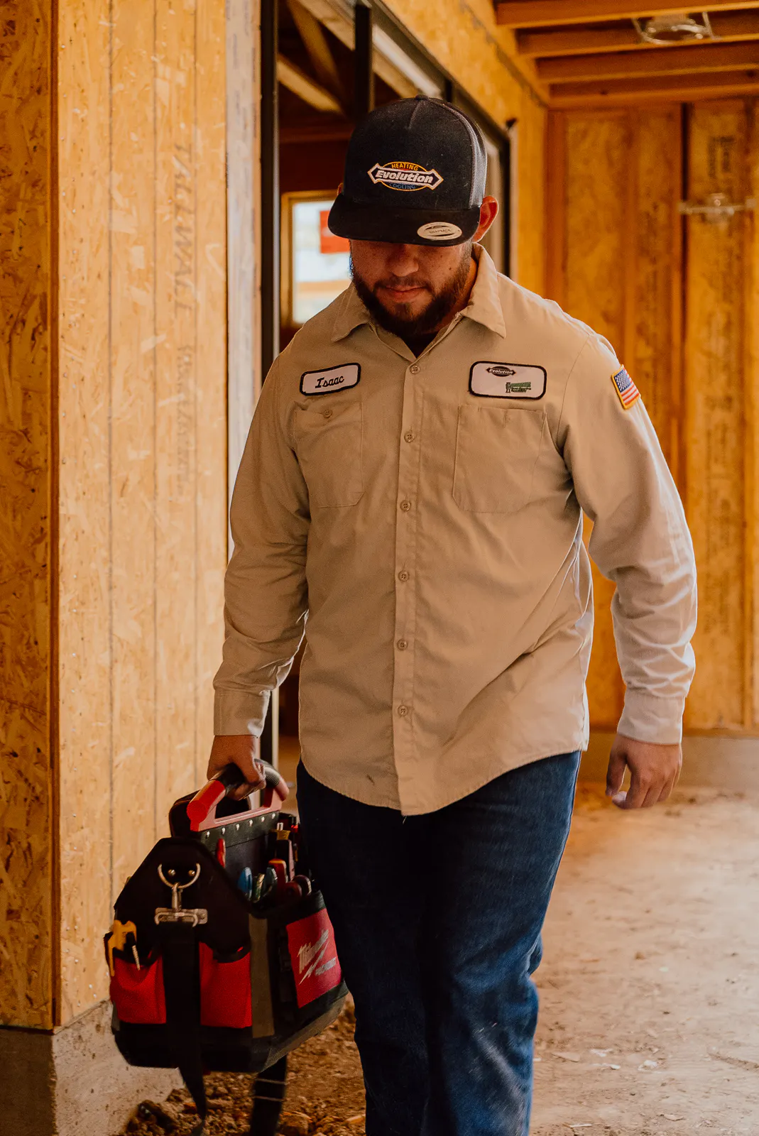 Male construction worker in beige shirt and cap carrying a tool bag inside a wooden framed building under construction.