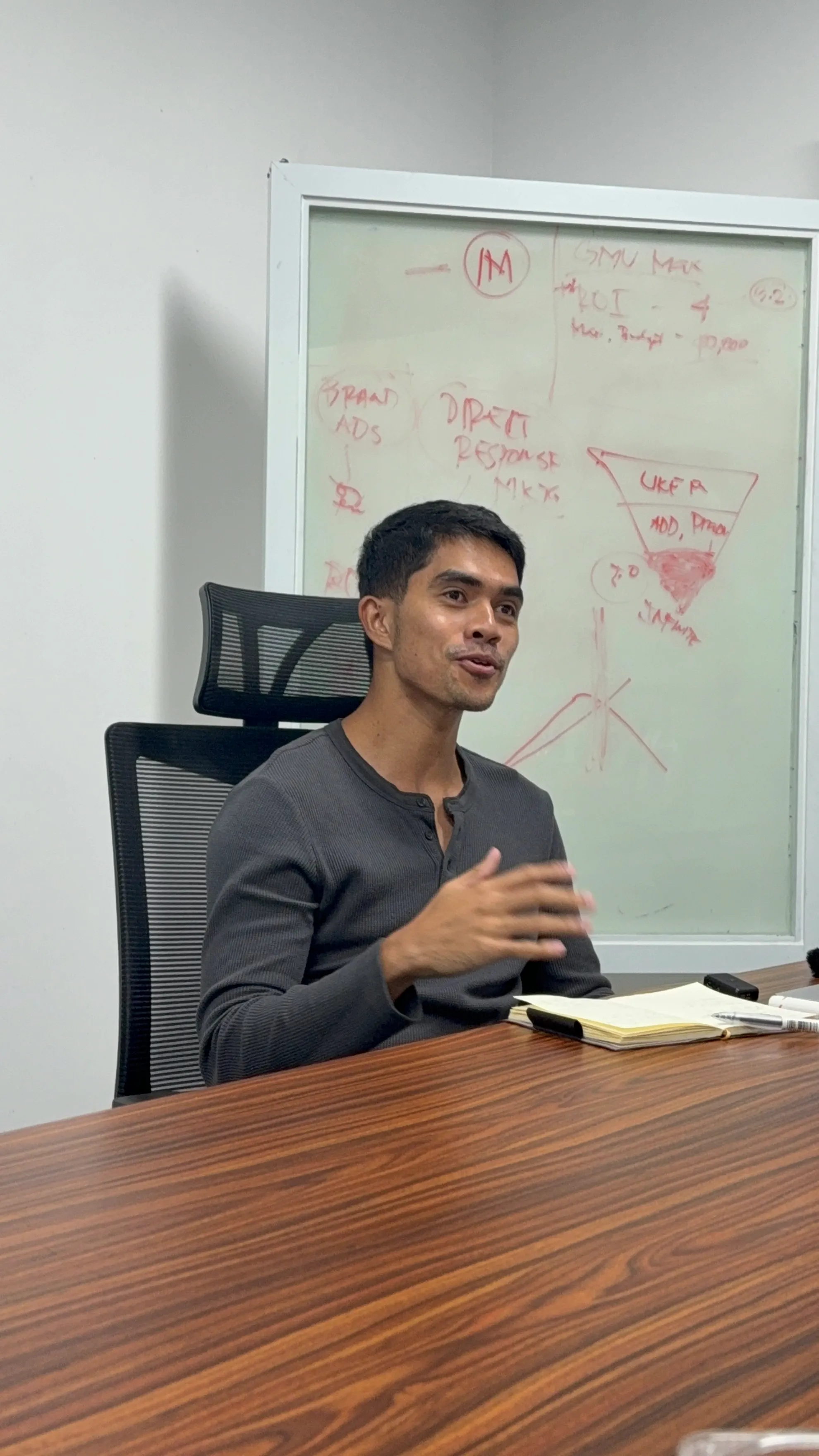 Man in a gray long-sleeve shirt speaking and gesturing with his right hand while seated at a wooden table with a notebook and pen, with a whiteboard containing red writing behind him.
