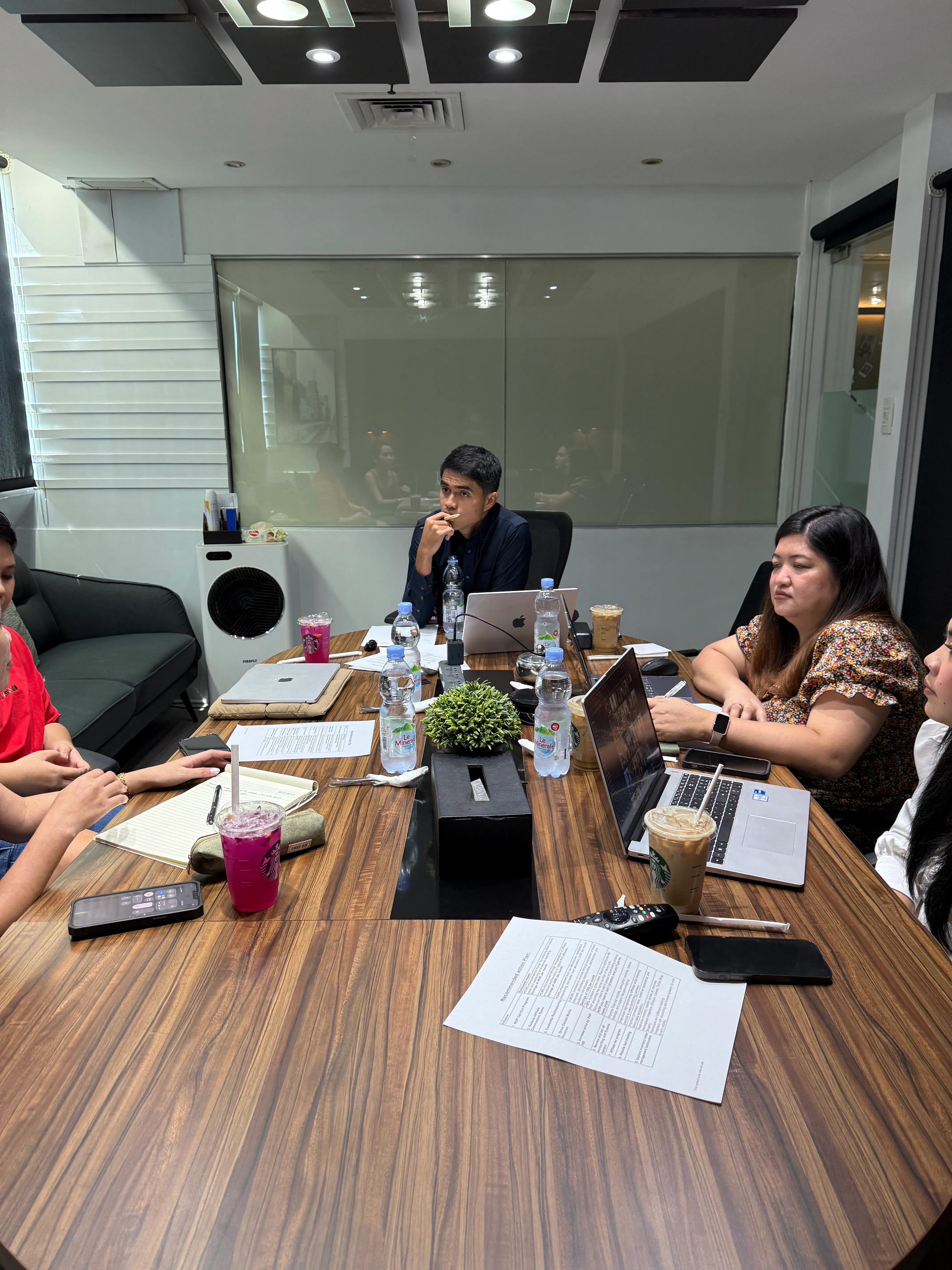 Four people seated around a wooden conference table with laptops, papers, drinks, and water bottles, in a meeting room with a glass partition wall.