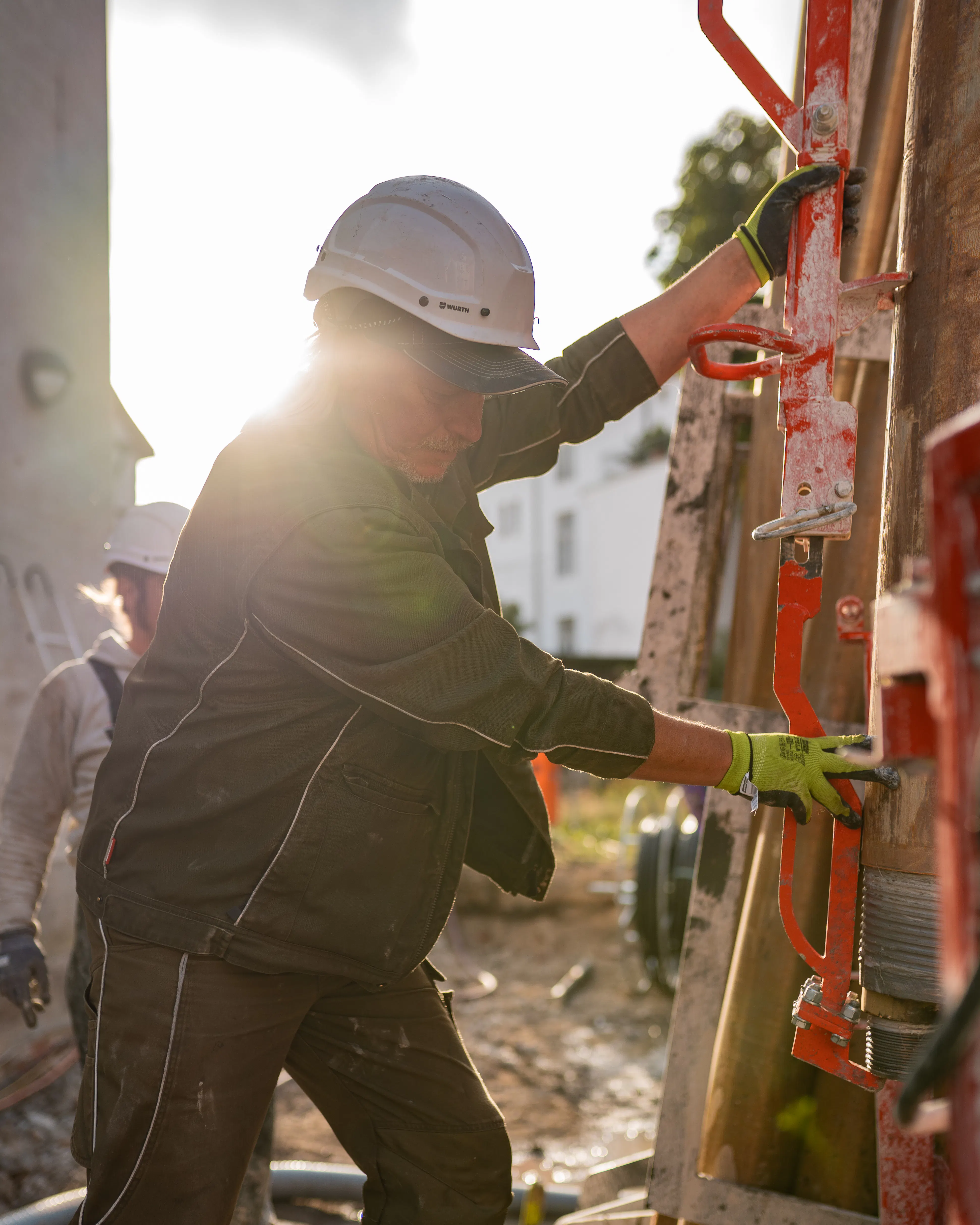Bauarbeiter mit Helm und Handschuhen bedient eine rote Stahlkonstruktion auf einer Baustelle bei Sonnenlicht.