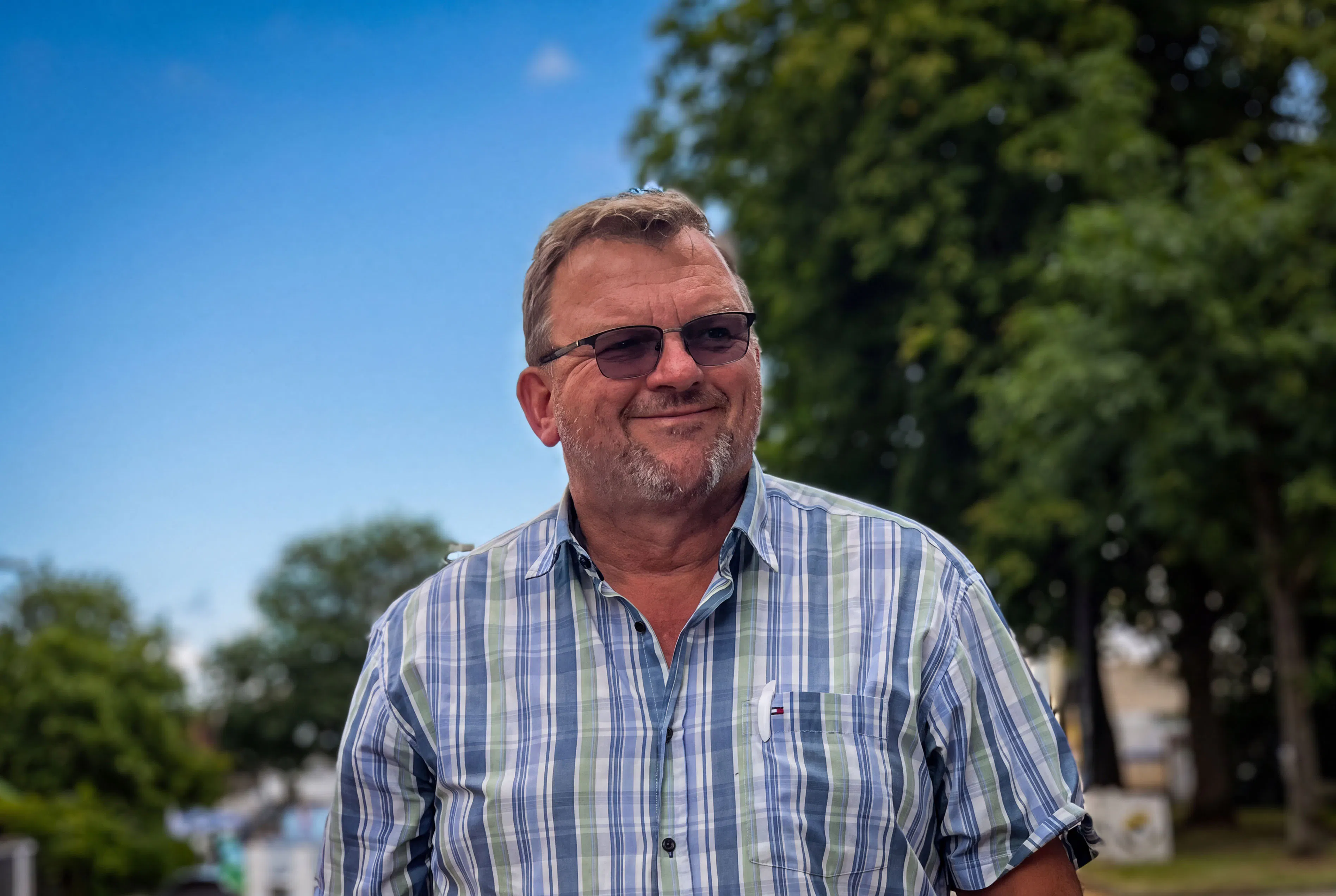 Mann mit Brille, kariertem Hemd und Stift in der Brusttasche steht draußen, im Hintergrund grüne Bäume und blauer Himmel.