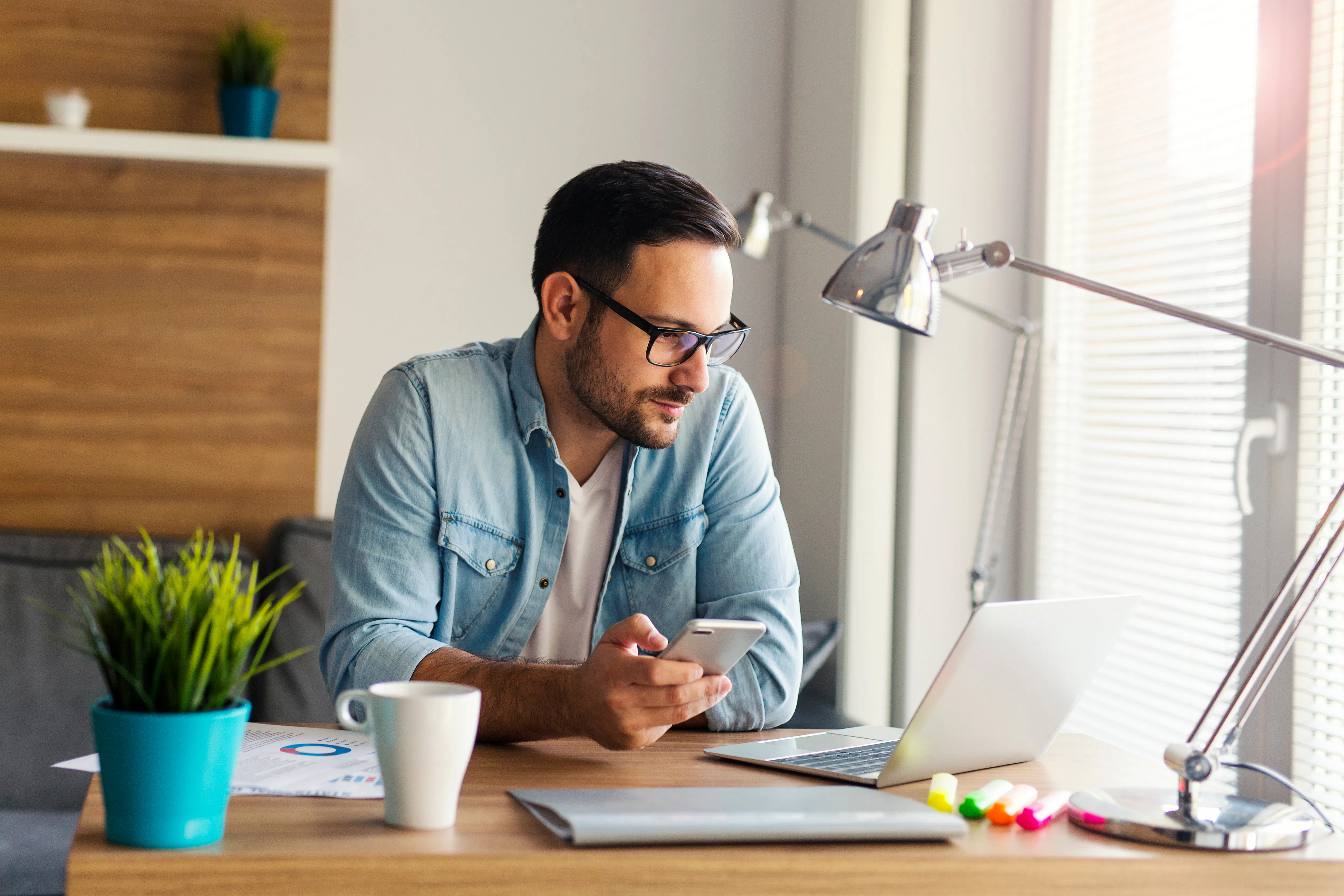 Mann mit Brille sitzt an einem Holztisch mit Laptop, Smartphone, Kaffeetasse, mehreren Markern und einem Pflanzentopf in einem hellen Raum mit Fenster und Schreibtischlampe.