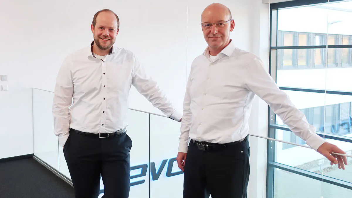 Two men in white shirts and black pants standing in a modern office space near a glass railing, one smiling with hands in pockets, the other with one arm resting on the railing.