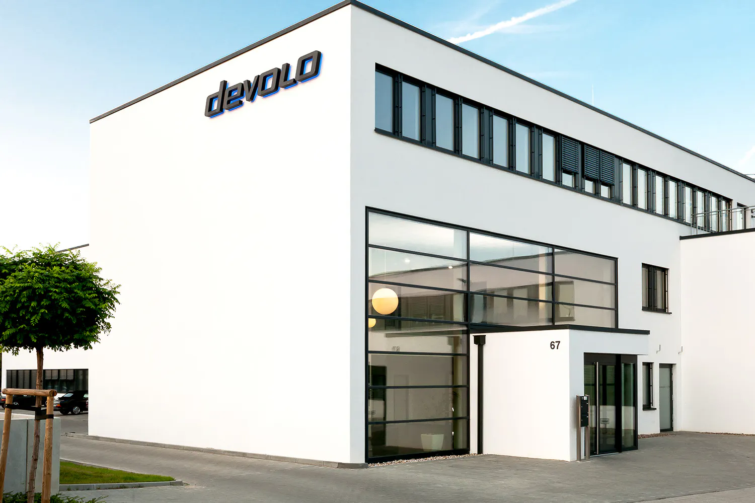 Modernes weißes Bürogebäude mit großen Glasfenstern und der schwarzen Aufschrift „devolo“ an der oberen Ecke, Hausnummer 67 am Eingang, kleiner Baum im Vordergrund.