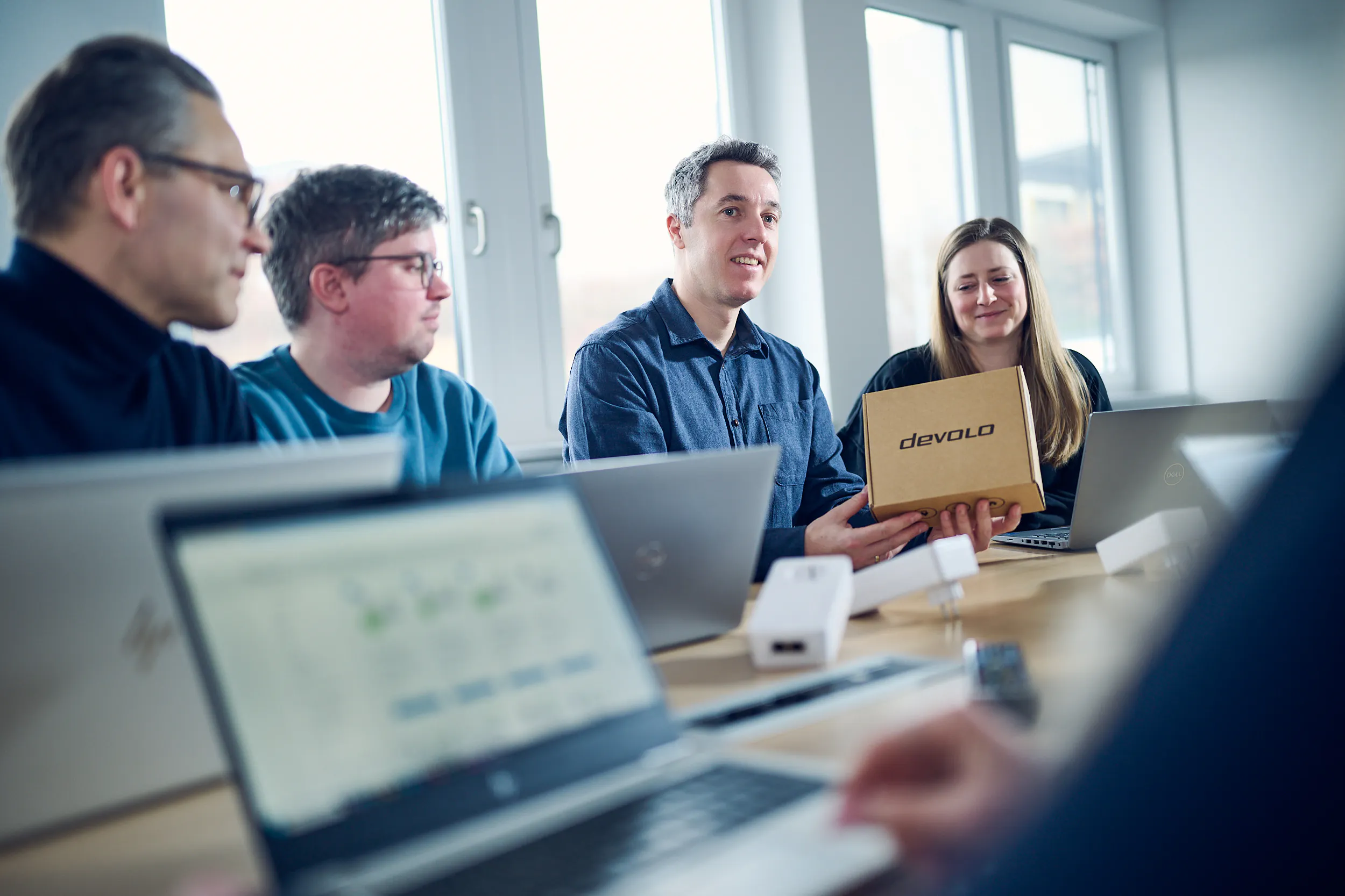 Person hält eine Kartonverpackung mit der Aufschrift "devolo", umgeben von anderen Personen mit Laptops in einem Besprechungsraum.