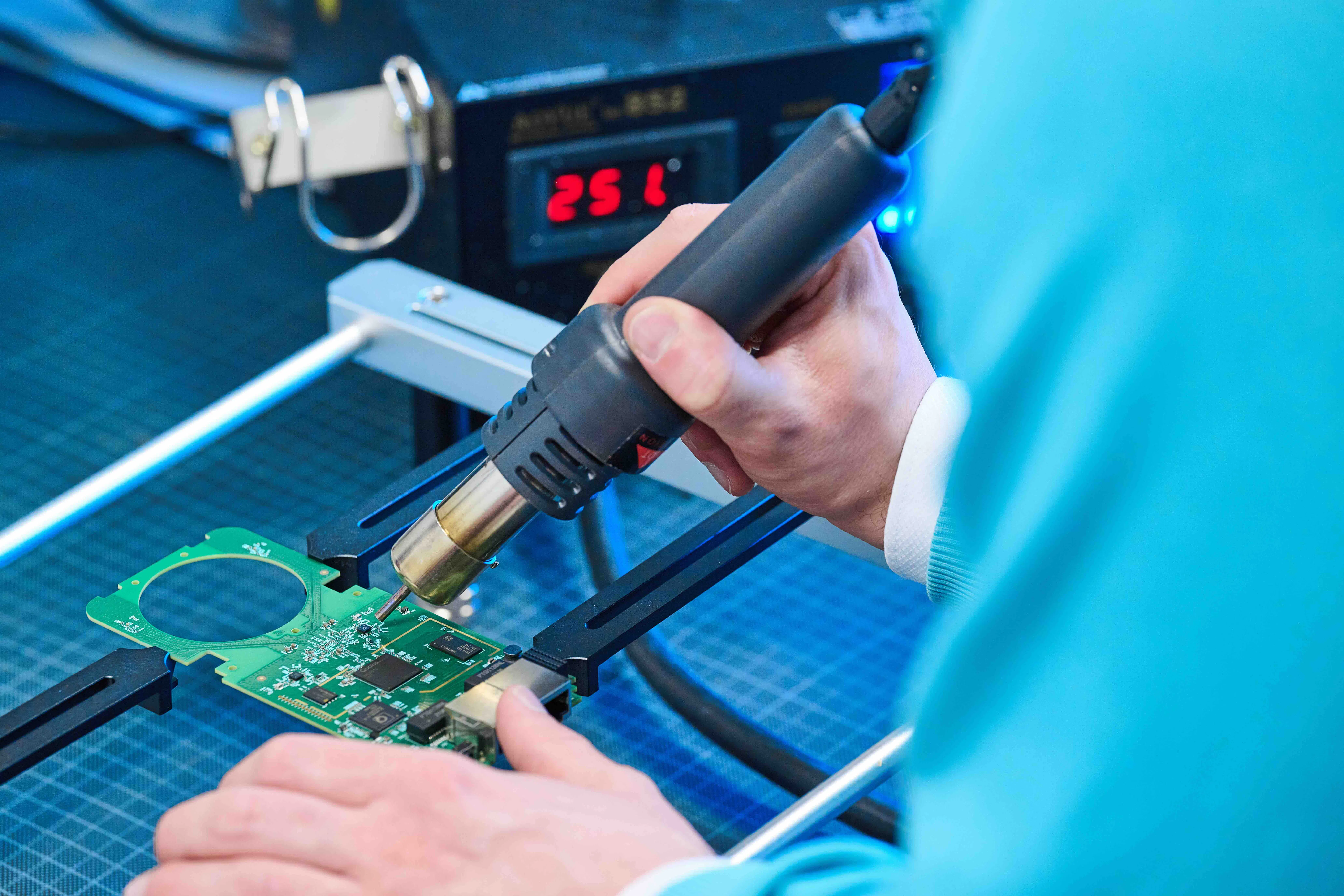 Hand holding a soldering iron repairing a green circuit board held in a fixture, with a digital display showing a temperature of 251 degrees in the background.