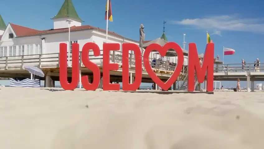 Große rote Buchstaben auf einem Sandstrand bilden das Wort "USEDOM" mit einem Herzsymbol anstelle des Buchstabens O, im Hintergrund eine Seebrücke und ein Gebäude mit grünem Dach.