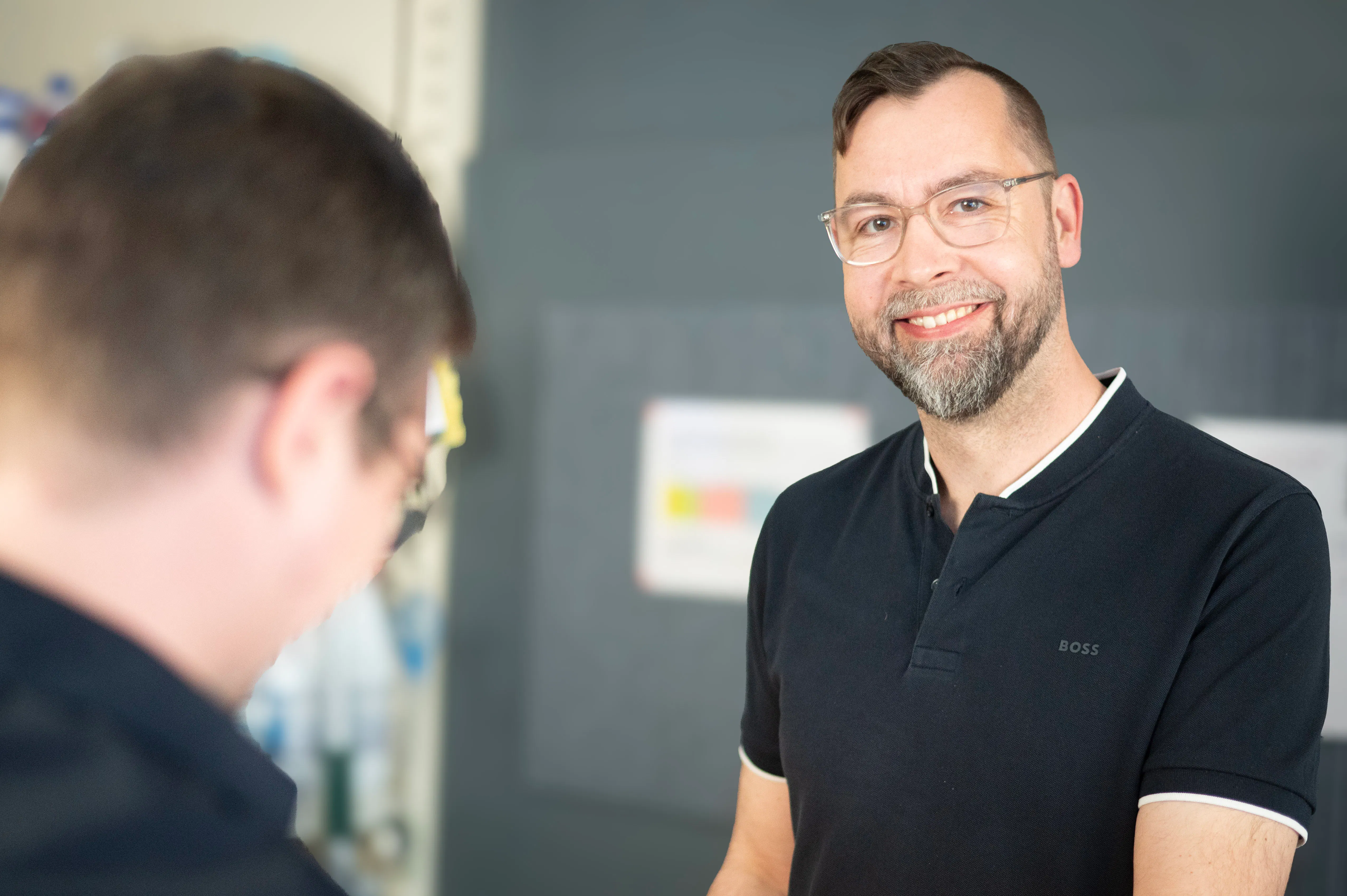 Zwei Männer im Gespräch, einer lächelnd und in den Fokus gerückt mit schwarzem Poloshirt und Brille, der andere unscharf im Vordergrund.