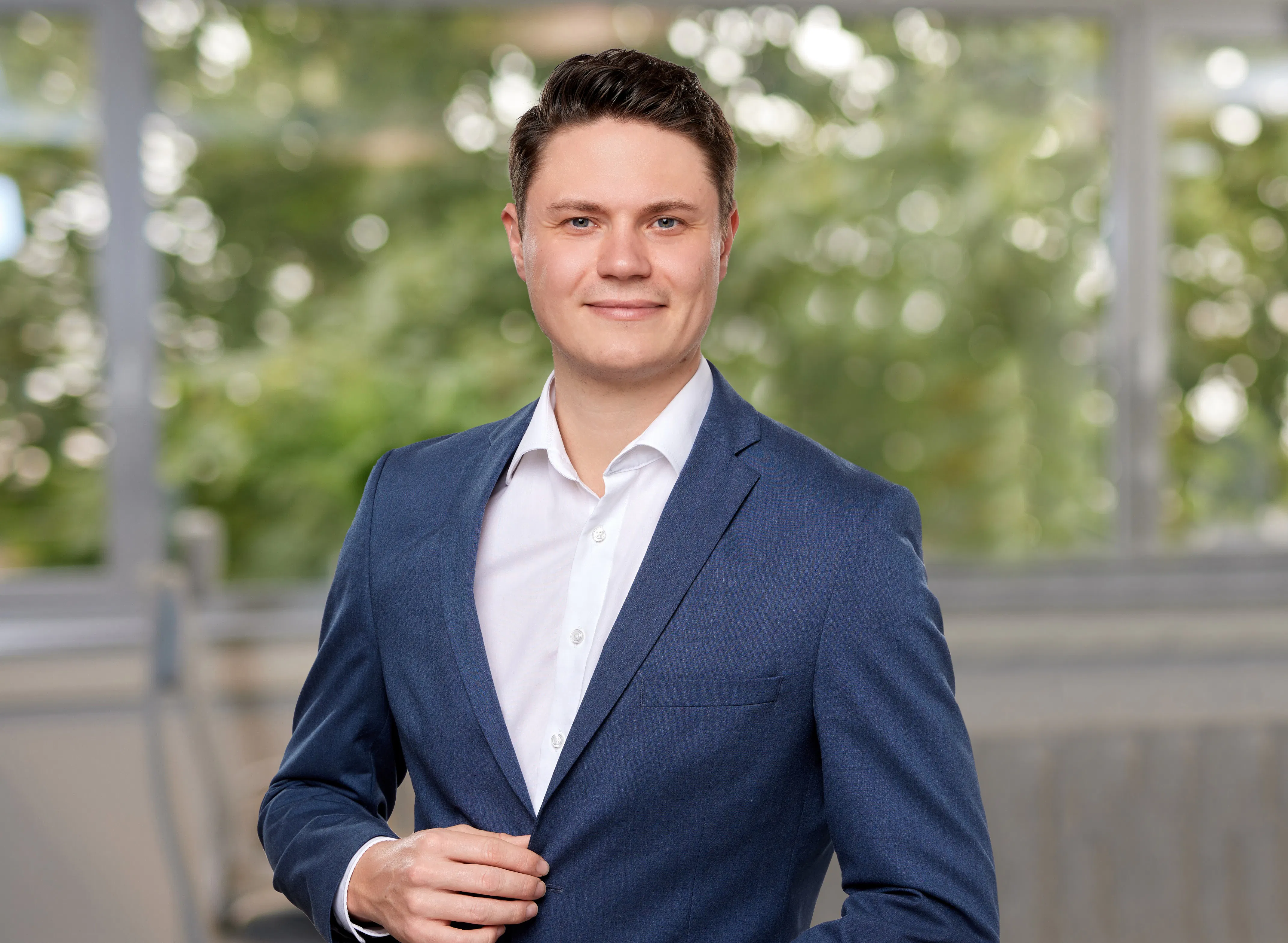 Junger Mann im blauen Anzug und weißem Hemd, der in einem hellen, unscharfen Innenraum in die Kamera lächelt.