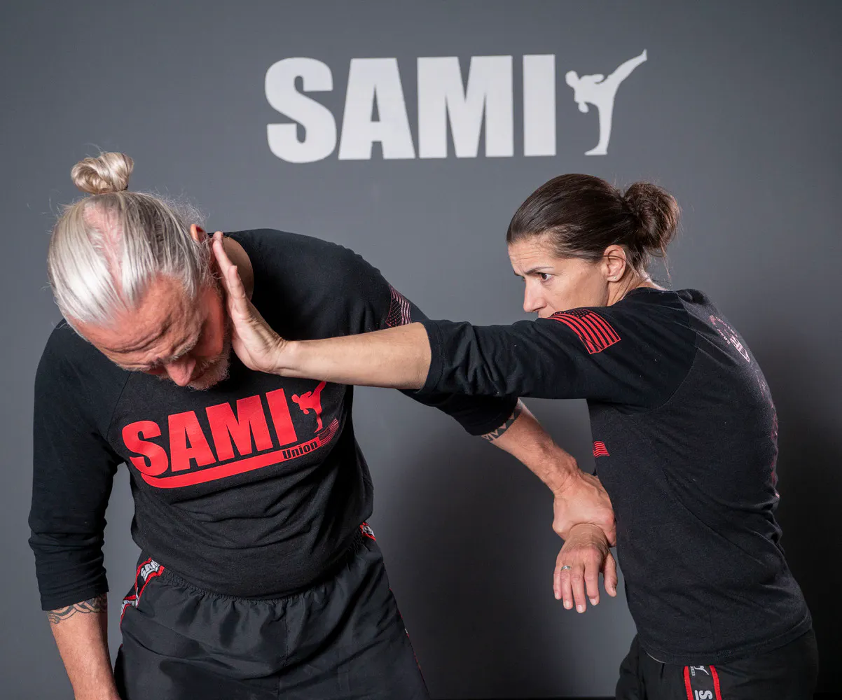 Frau übt eine Selbstverteidigungstechnik mit einem Mann in Trainingskleidung vor einer Wand mit dem Schriftzug "SAMI".