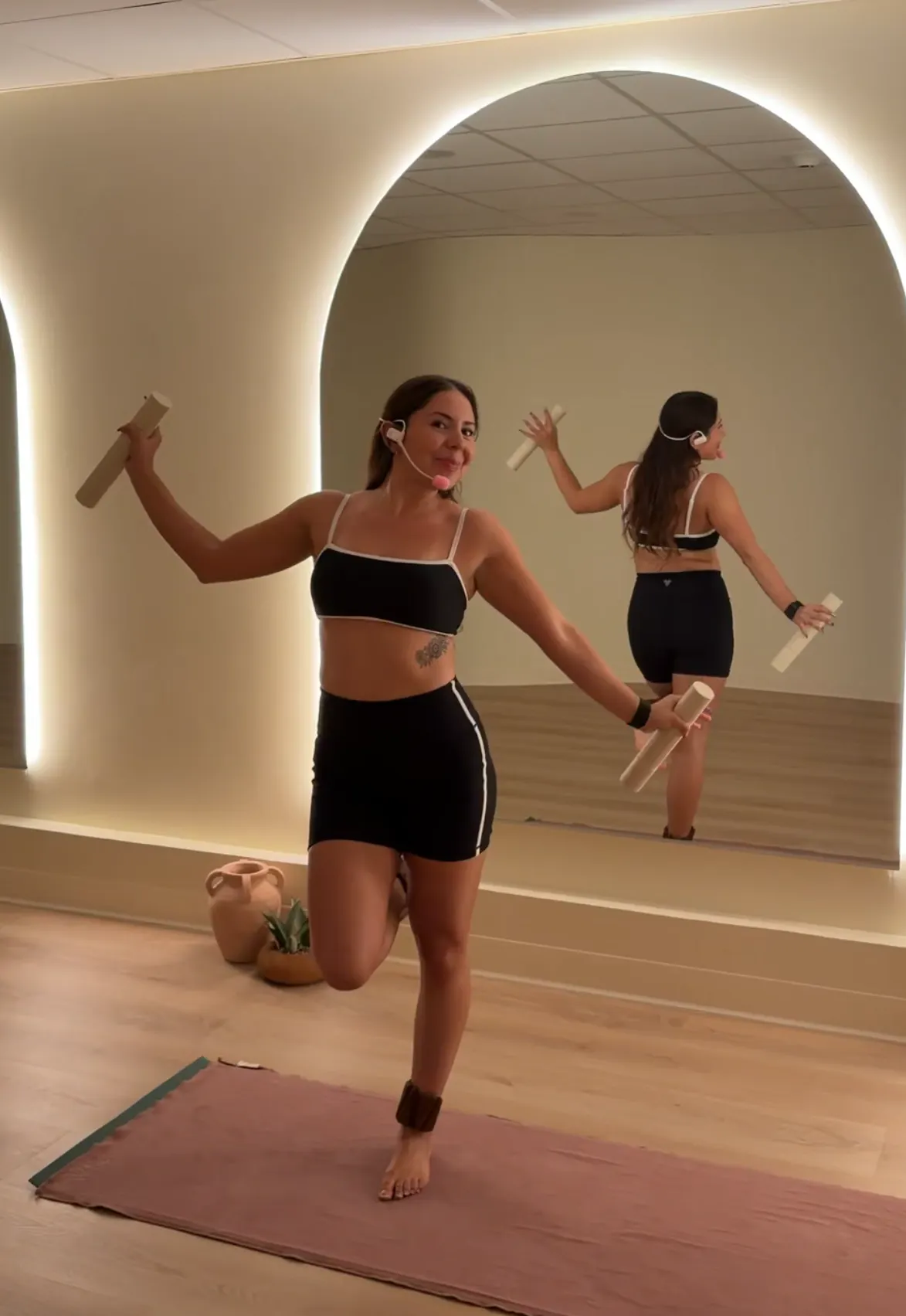 Woman in black workout outfit balancing on one leg on a mat, holding two light sticks, wearing a headset, standing in front of a large illuminated mirror in a studio.