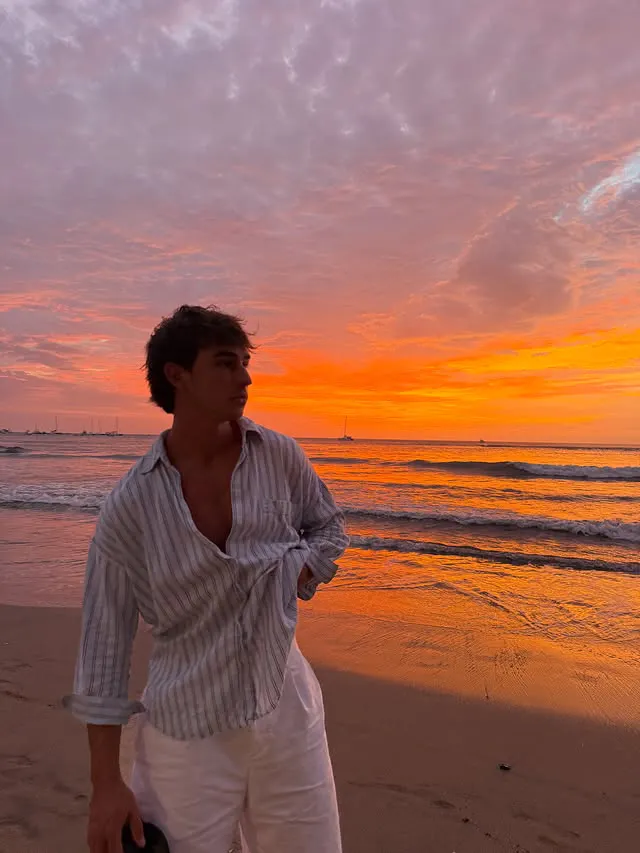 Young man wearing a striped shirt and white pants standing on a beach during a vibrant orange and purple sunset with calm waves and sailboats in the distance.