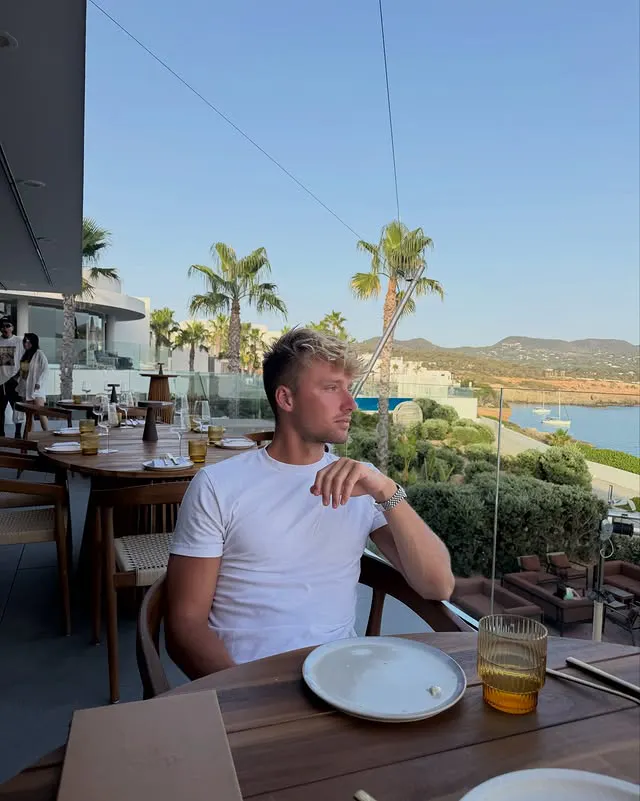 Young man in a white t-shirt sitting at a restaurant table outdoors with empty plates and a glass, facing a view of palm trees, hills, and water under a clear sky.