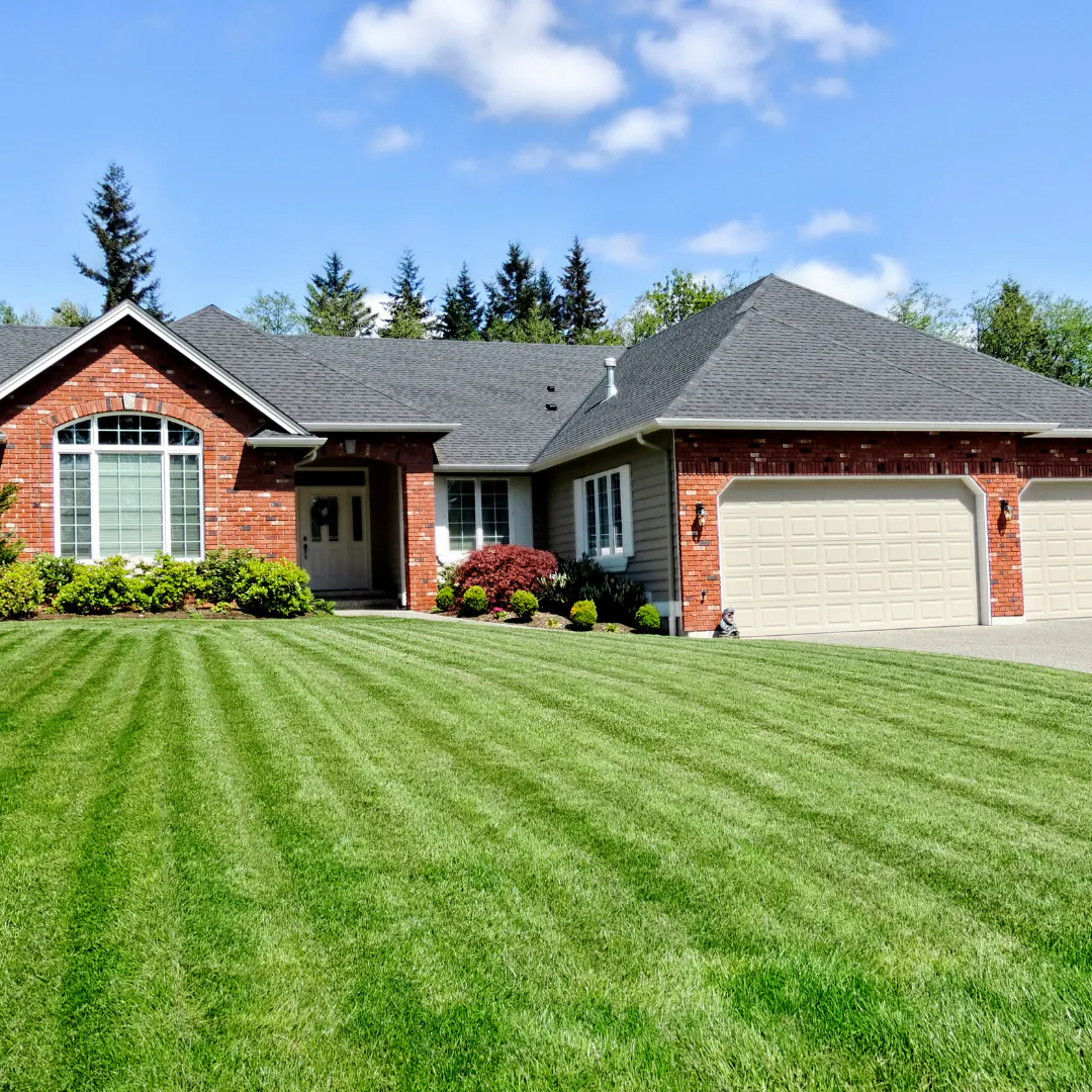 Single-story brick house with a large, well-maintained green lawn, three-car garage, and surrounding shrubs under a partly cloudy blue sky.
