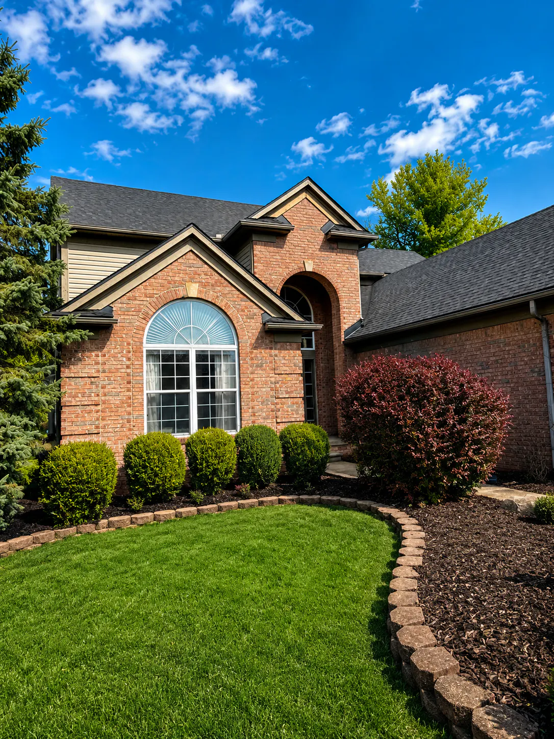 Brick house with arched window, manicured green lawn, and shrubs under a blue sky with scattered clouds.
