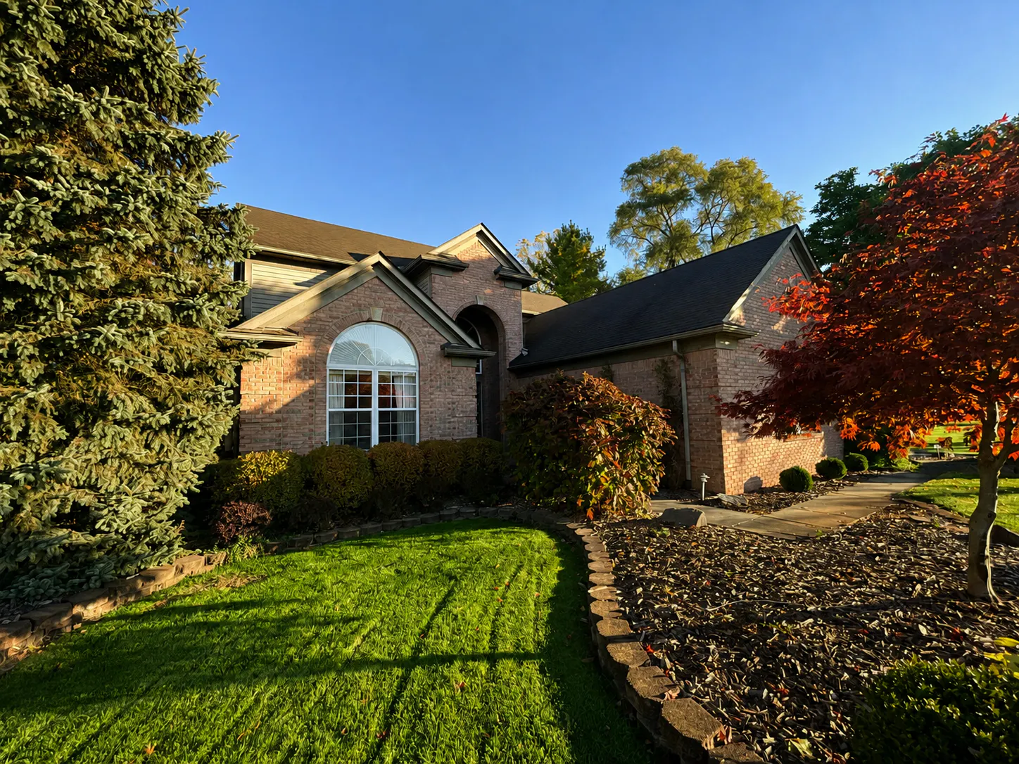 Brick house with arched window and landscaped front yard featuring green lawn, evergreen trees, shrubs, and a small red-leaved tree under a clear blue sky.