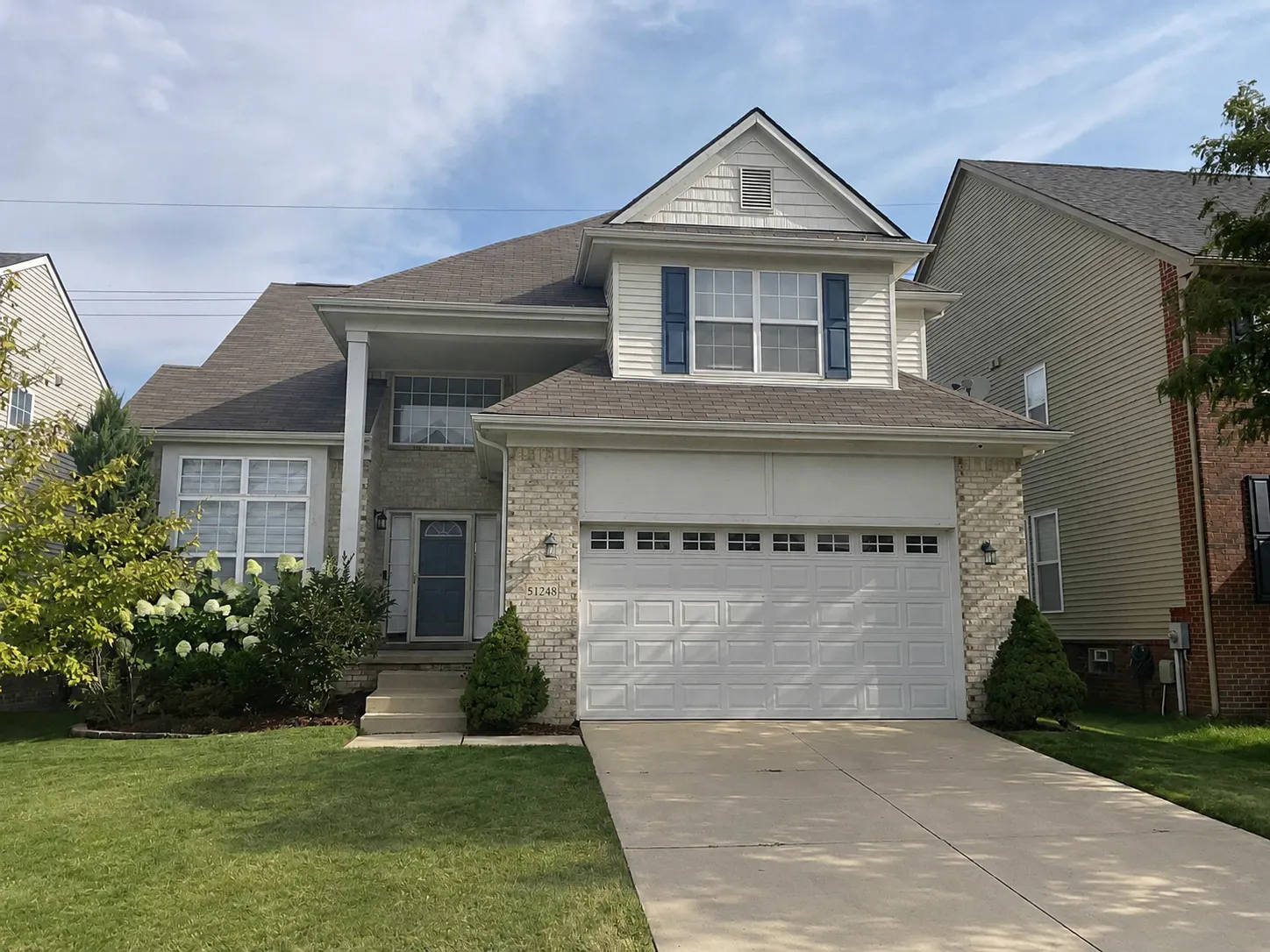 Two-story suburban house with a double garage, brick and siding exterior, blue front door, and a front lawn with shrubs and a concrete driveway.