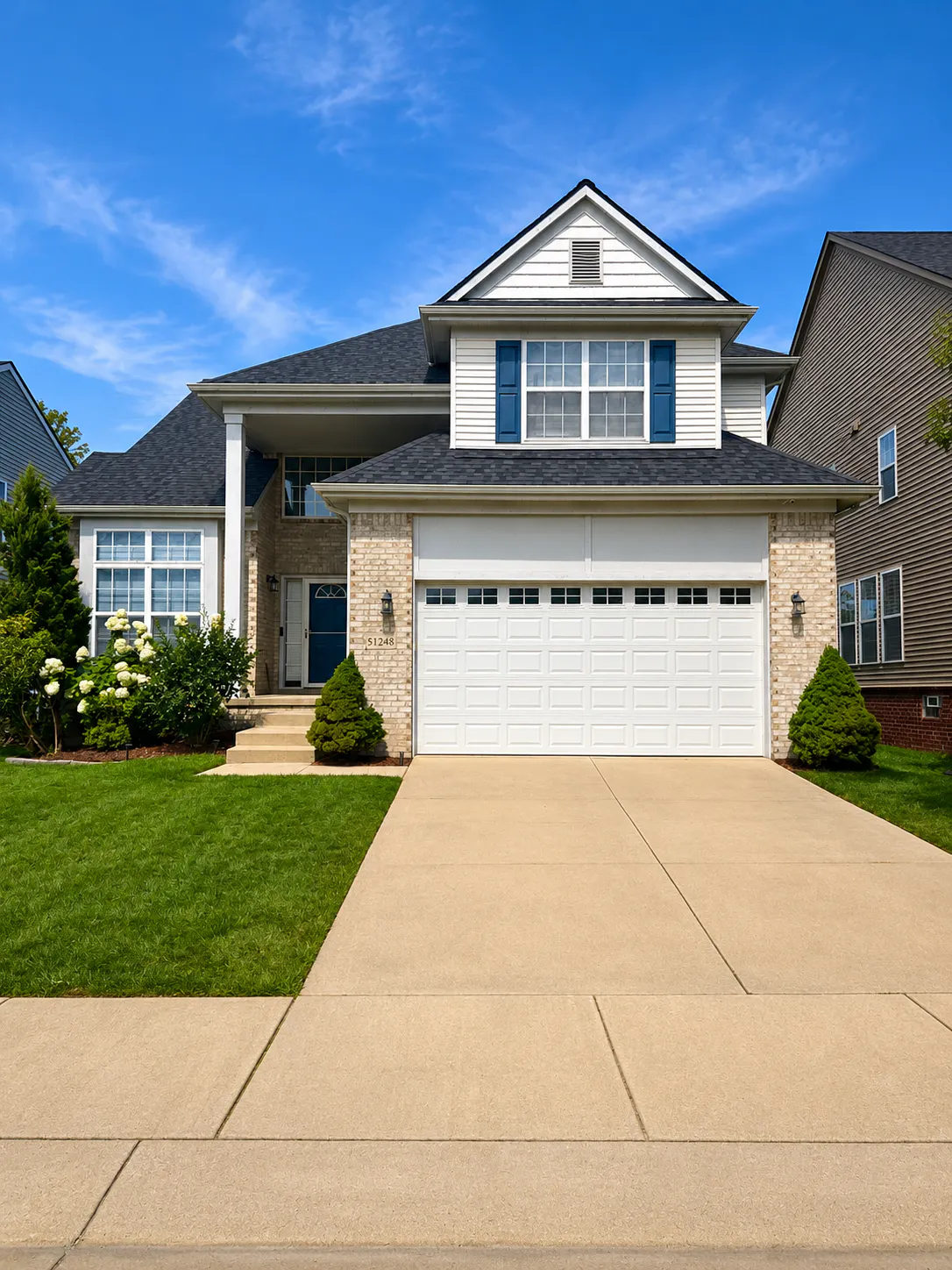 Two-story suburban house with beige brick facade, white garage door, blue shutters, small lawn, and clear blue sky above.