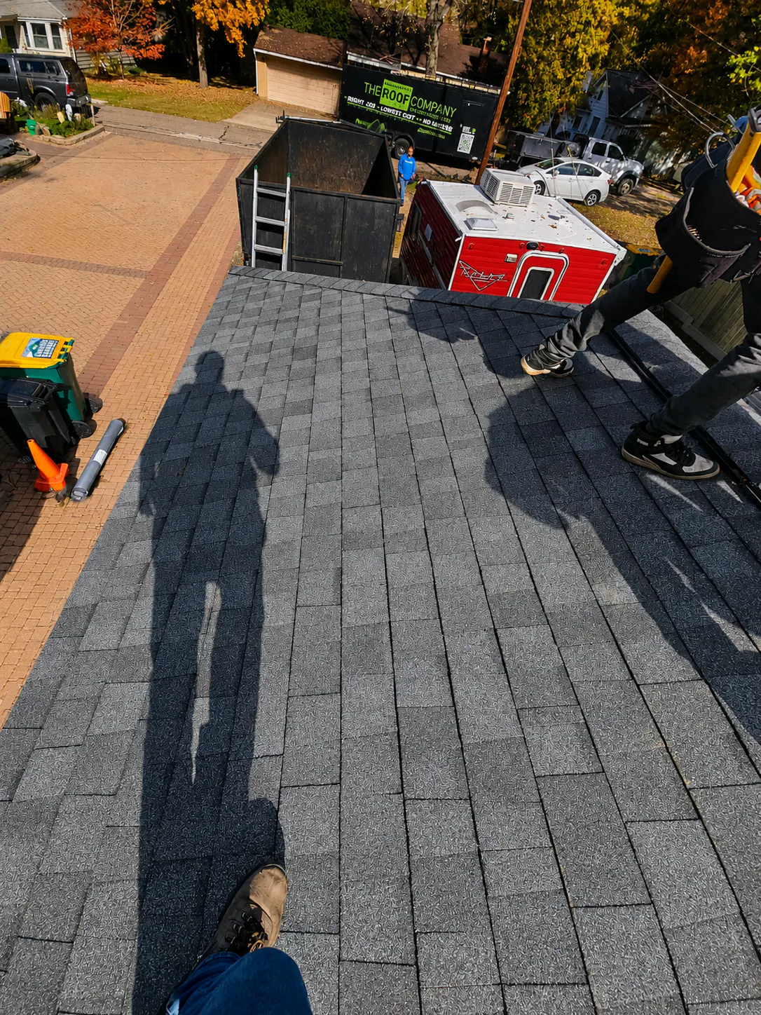 Footwear and shadows of two people standing on a rooftop with gray shingles, with a street, vehicles, a dumpster, and autumn foliage visible below.