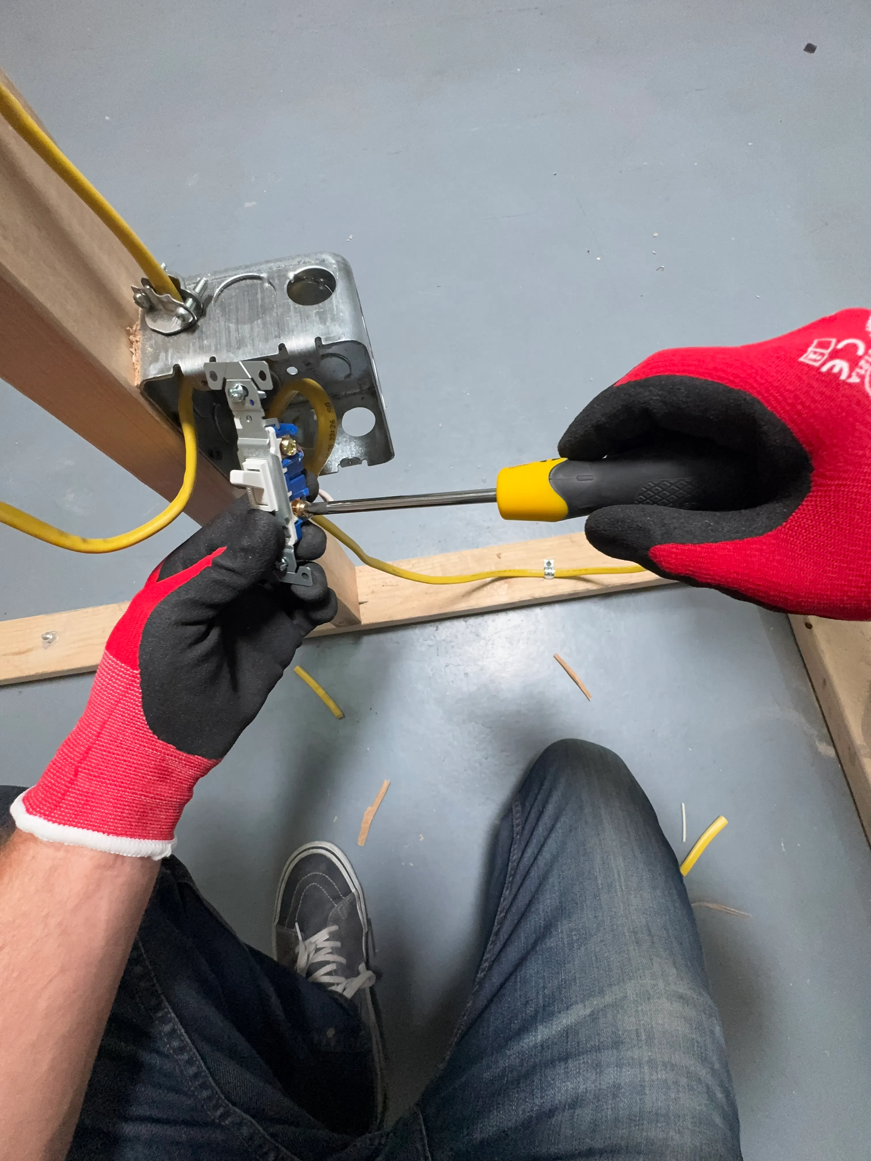 Hands wearing red and black gloves install an electrical switch into a metal junction box with a yellow screwdriver, with yellow electrical wires connected inside.