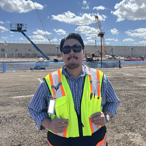 Man wearing sunglasses and a high-visibility safety vest standing on a construction site with cranes and industrial equipment in the background under a partly cloudy sky.