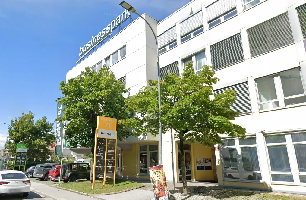 Firmengebäude mit Geschäftsschildern, darunter die Aufschrift "businesspark", auf einem sonnigen Straßenzug mit Bäumen und parkenden Autos.