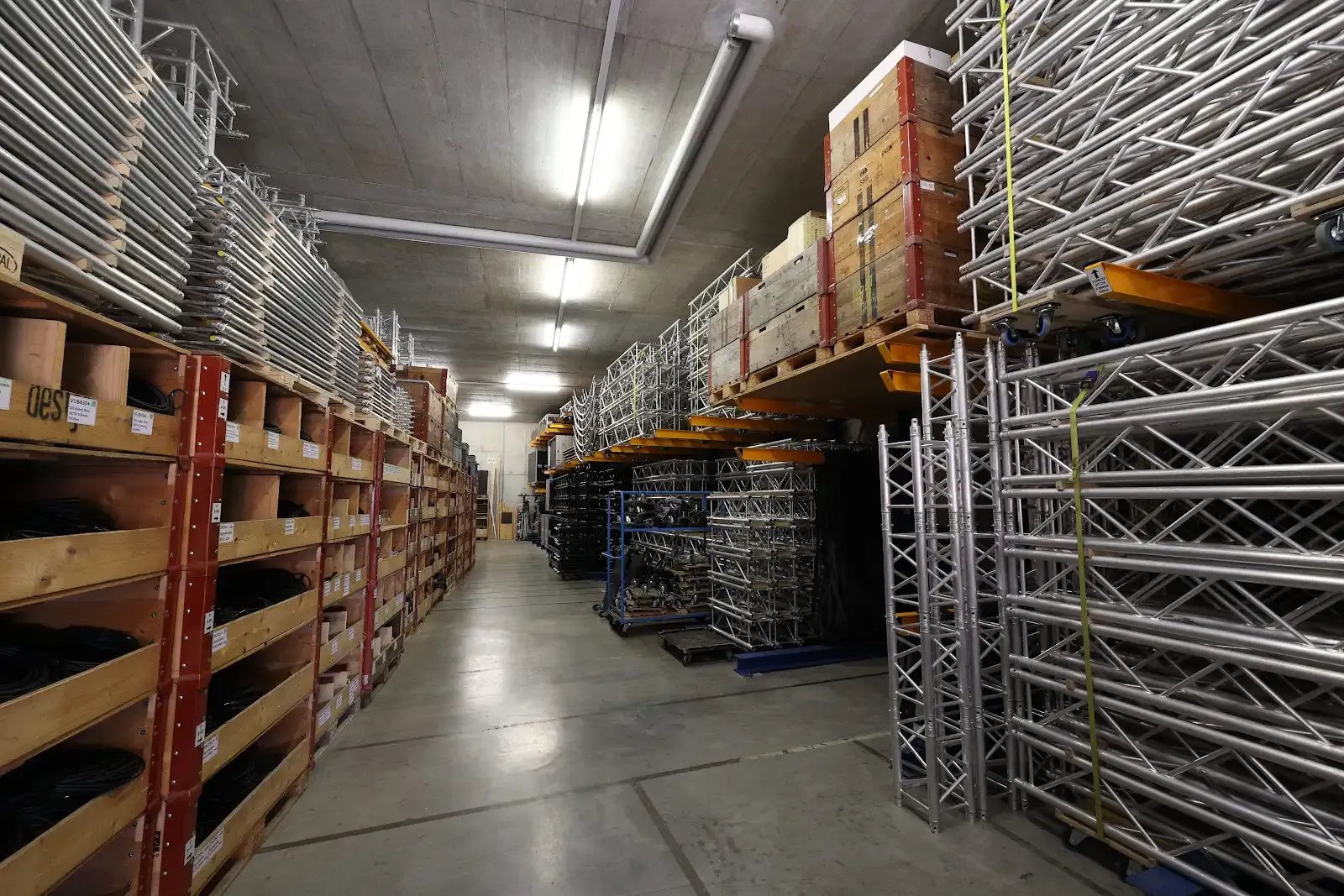 Lagerhalle mit Metallgestellen und Holzkisten, Aufbewahrung von Bühnen- oder Eventausrüstung auf Regalen und dem Boden, beleuchteter Raum mit Betonboden und -decke.