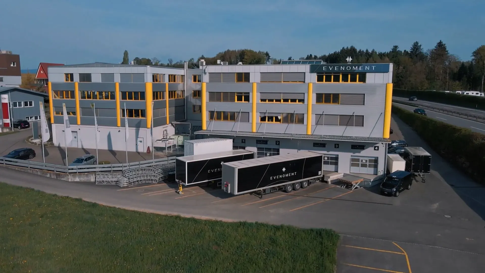 Modernes graues Bürogebäude mit gelben Säulen, zwei schwarzen Lastwagen mit "Evenoment"-Schriftzug und mehreren parkenden Autos auf einem Industrieparkplatz.