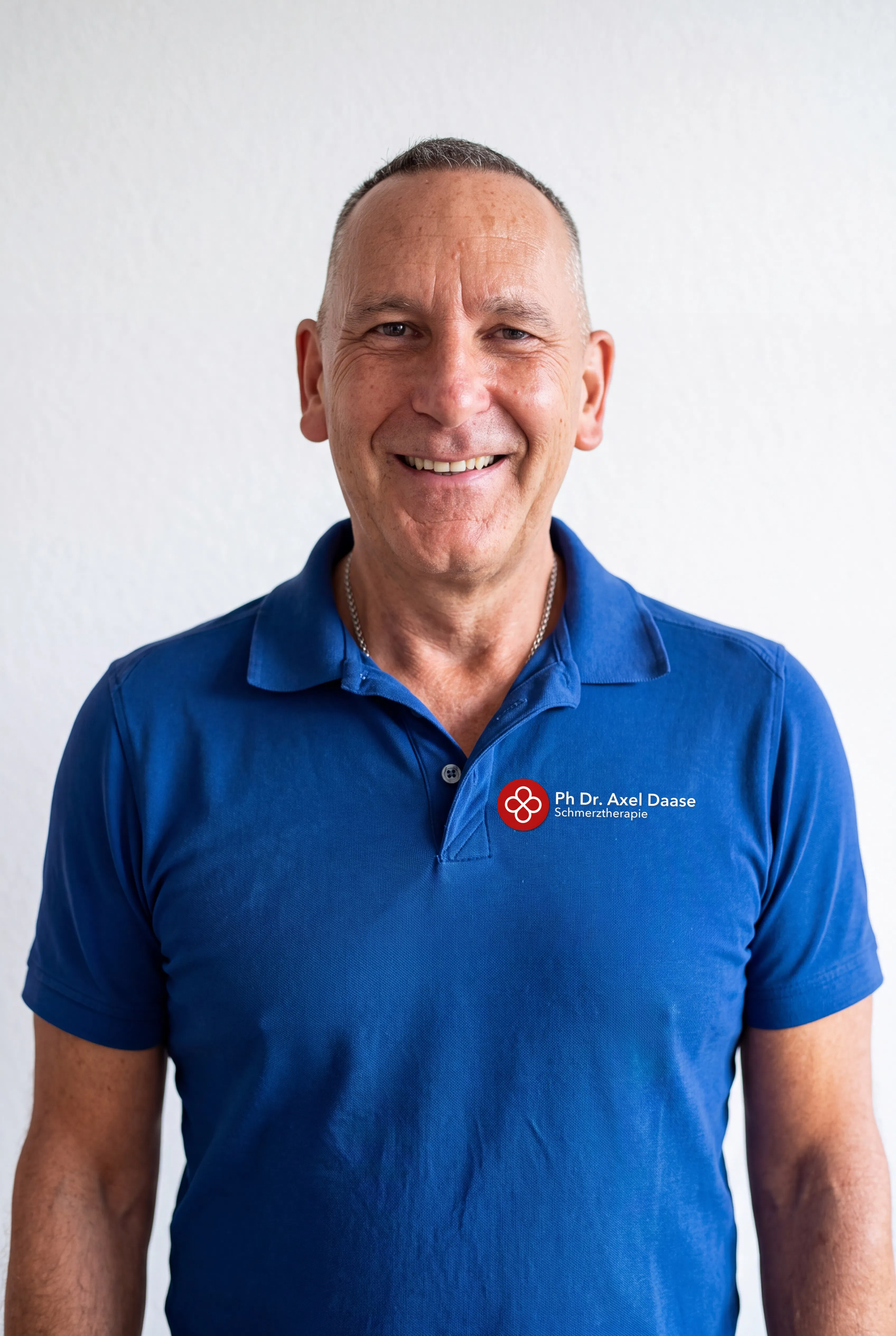 Lächelnder Mann mit kurzem grauem Haar trägt ein blaues Poloshirt mit Logo und Schriftzug „Ph Dr. Axel Daase Schmerztherapie“ vor weißem Hintergrund.