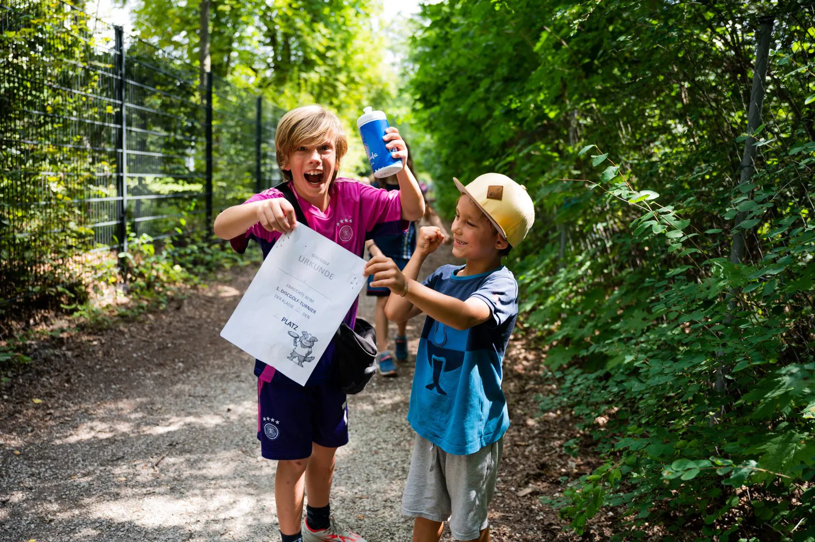 Zwei Jungen auf einem Waldweg, einer hält eine Urkunde und eine Trinkflasche, beide zeigen freudige Gesten.