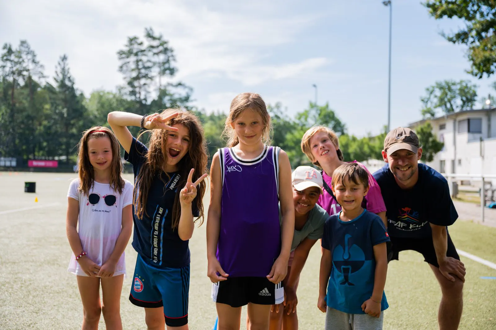 Group of six children and one adult standing on a sports field, some smiling and one girl making peace signs with her fingers.