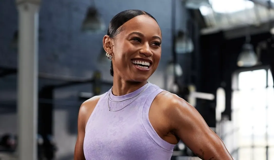 Smiling woman in a lavender sleeveless athletic top standing in a gym with gym equipment and large windows in the background.