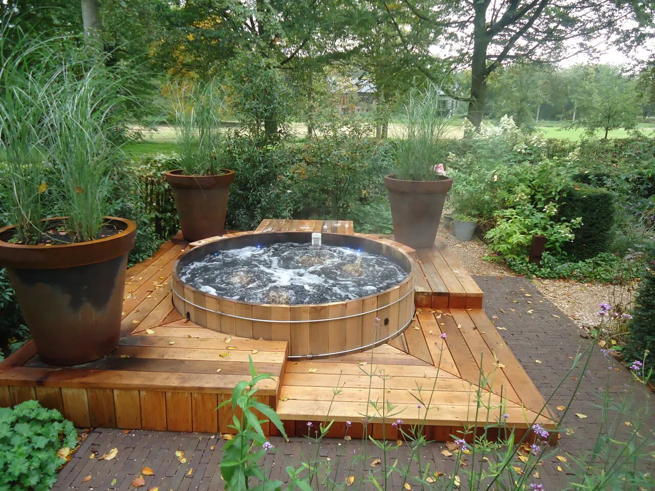 Round wooden hot tub with bubbling water on wooden deck surrounded by large potted plants and garden greenery in an outdoor setting.
