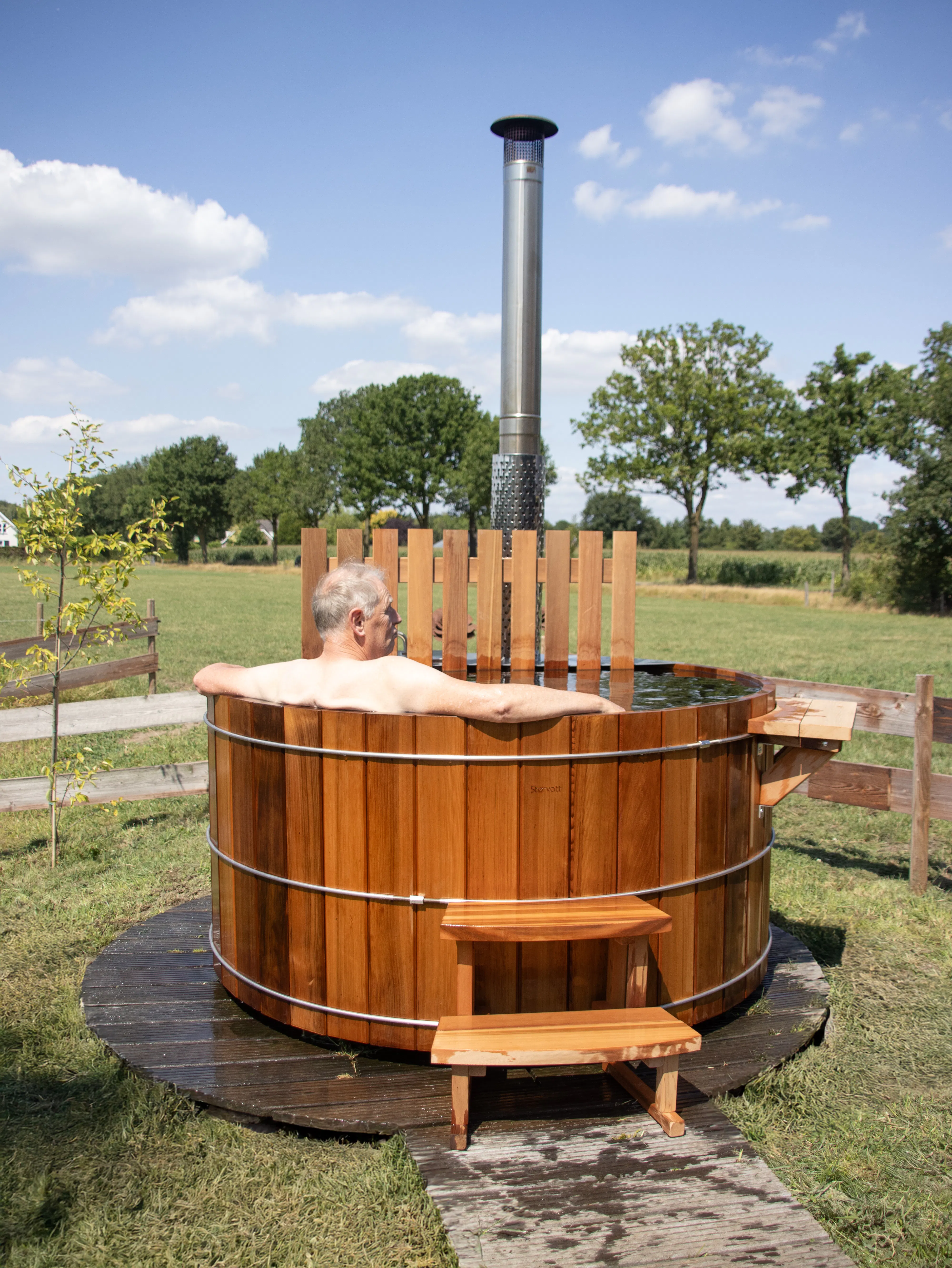 Man sitting in a round wooden hot tub with a metal chimney, placed outdoors on grass with trees and a blue sky in the background.