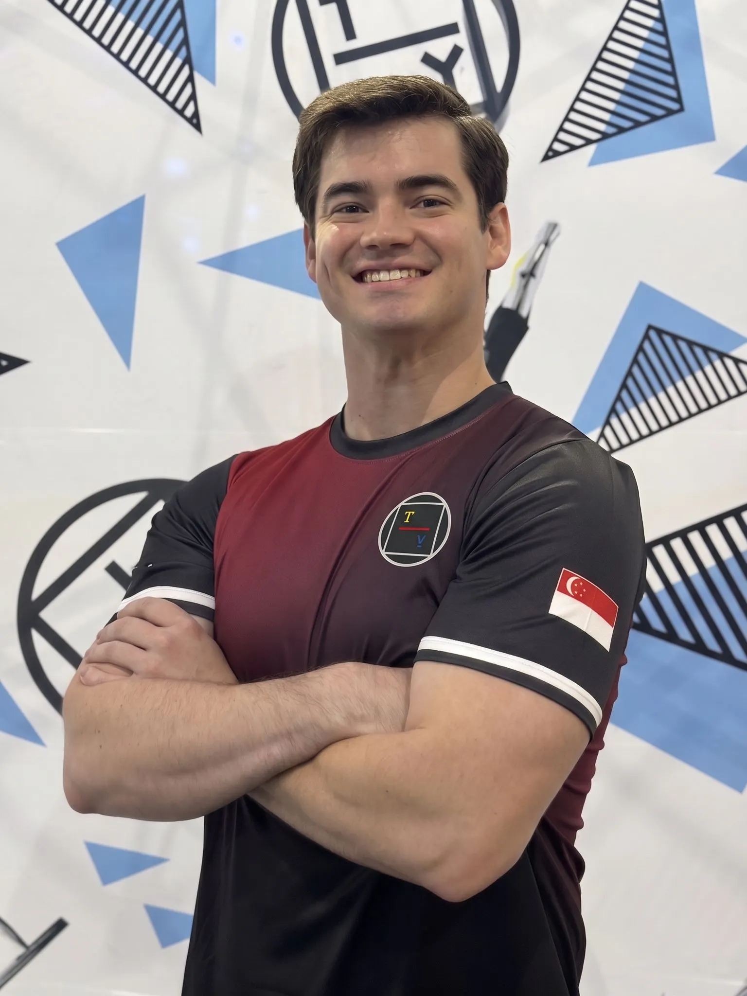 Smiling man with folded arms wearing a maroon and black shirt with a Singapore flag patch on the sleeve and a circular logo on the chest, standing in front of a patterned background.