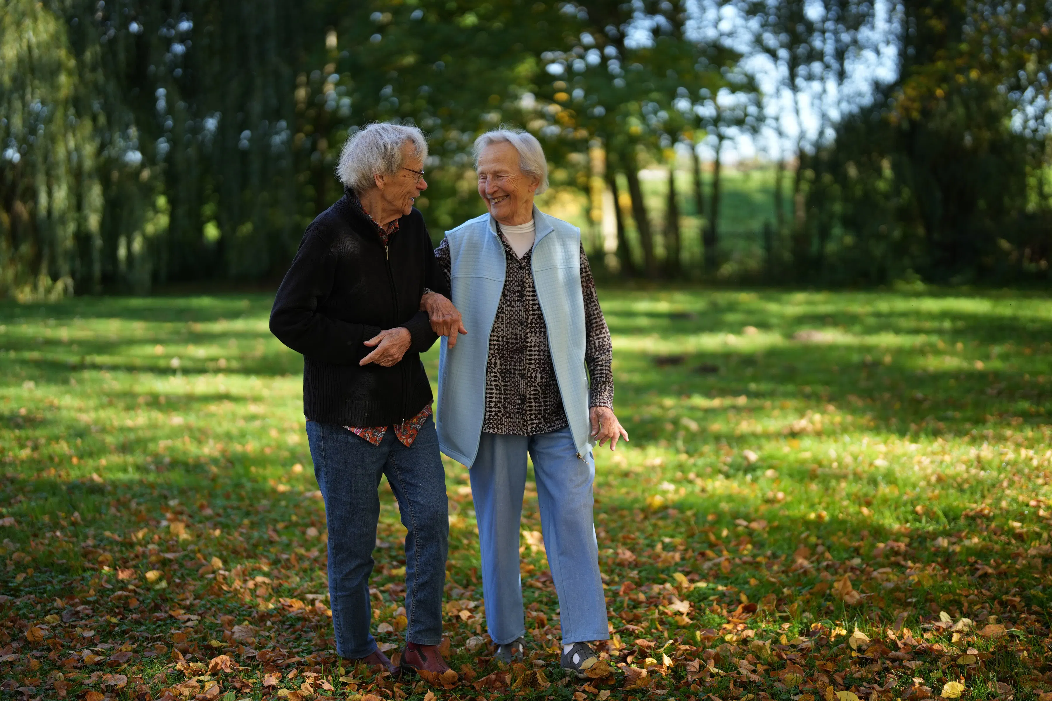 Zwei ältere Frauen, die Arm in Arm im Herbstlaub auf einer Wiese vor Bäumen spazieren gehen und lächeln.