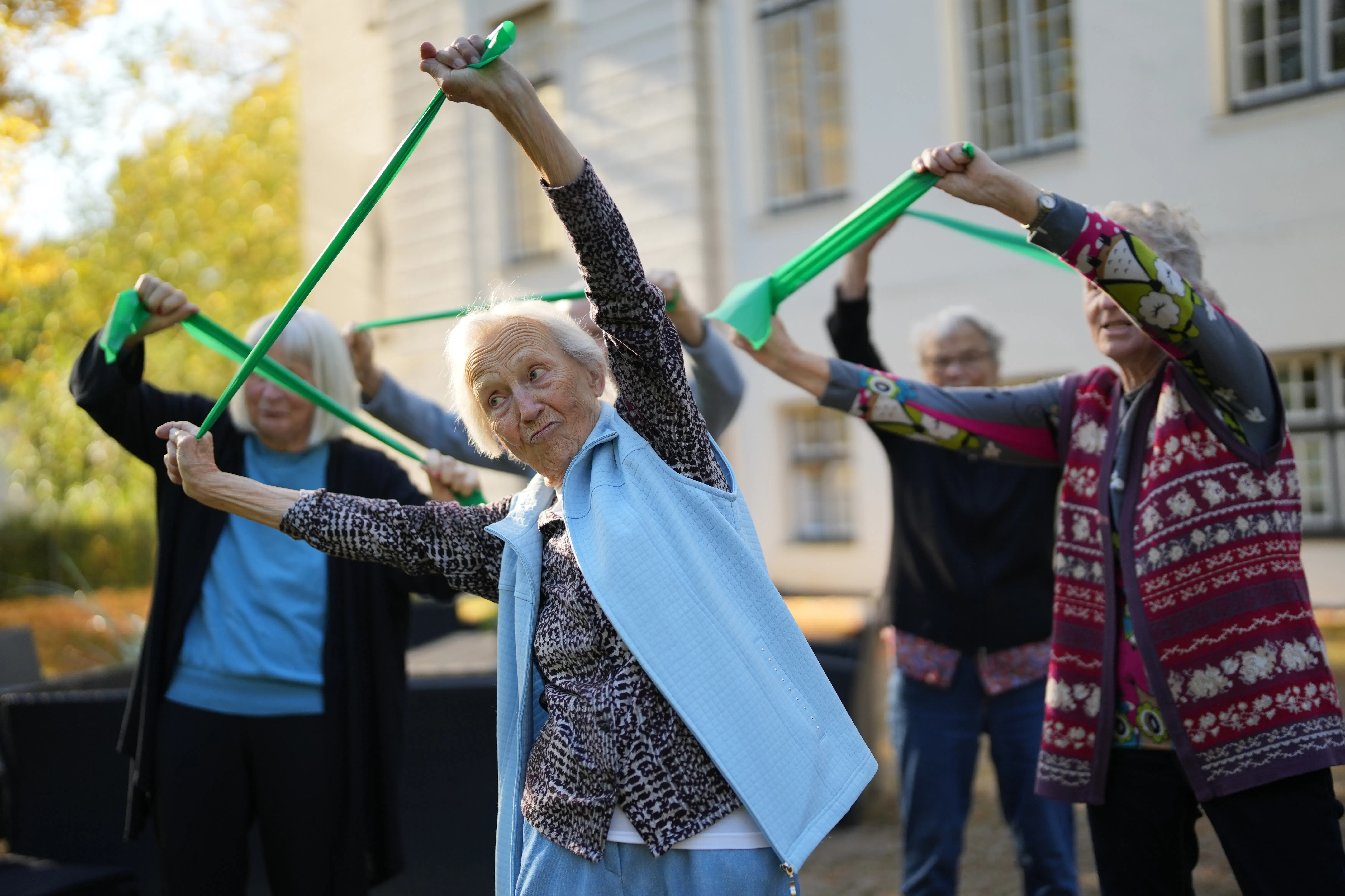 Ältere Menschen machen gemeinsam Dehnungsübungen mit grünen Fitnessbändern im Freien vor einem Gebäude.