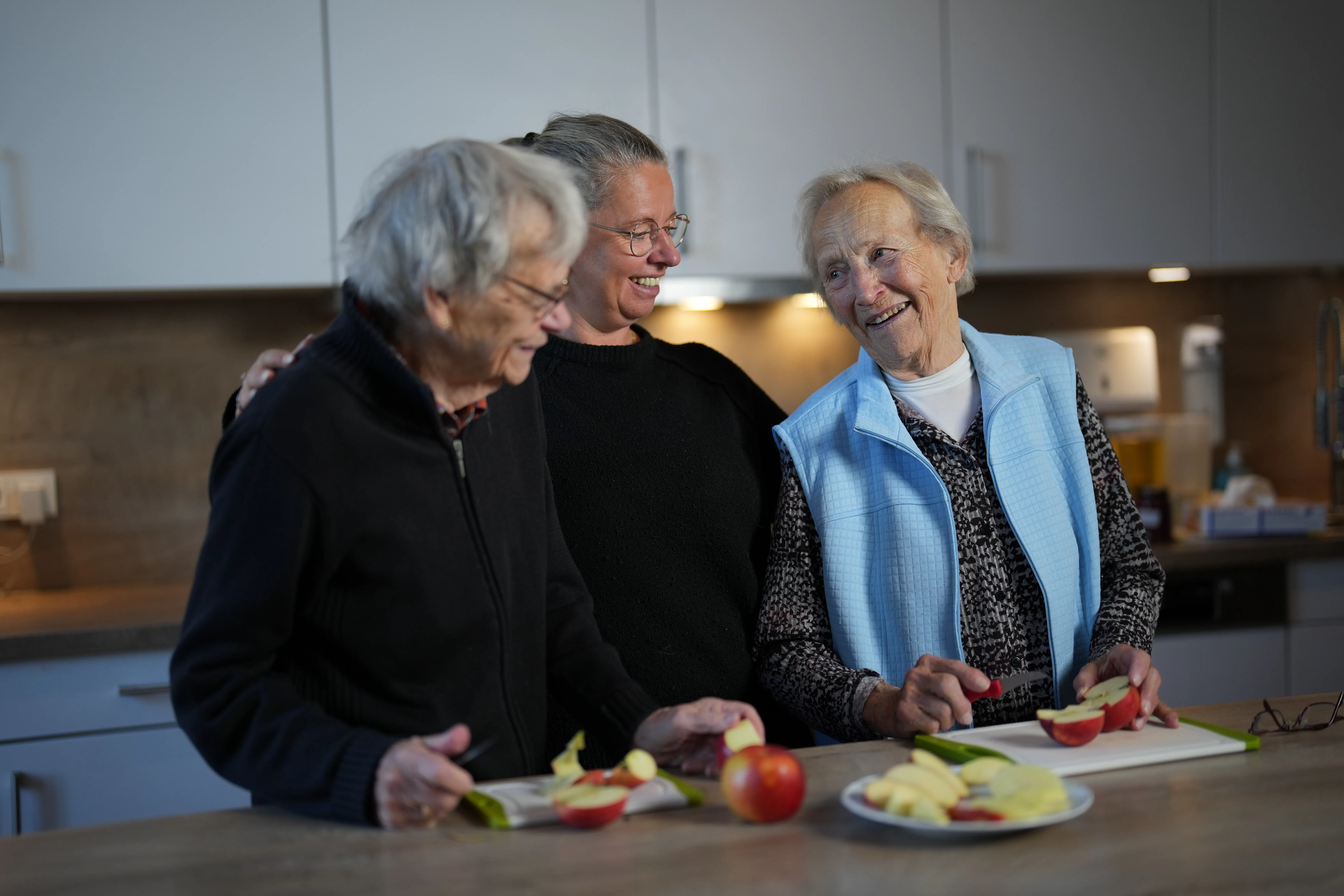 Drei Frauen beim Apfelschneiden in einer Küche, zwei ältere Damen schneiden Äpfel, die Frau in der Mitte lacht und legt ihren Arm um eine der älteren Damen.