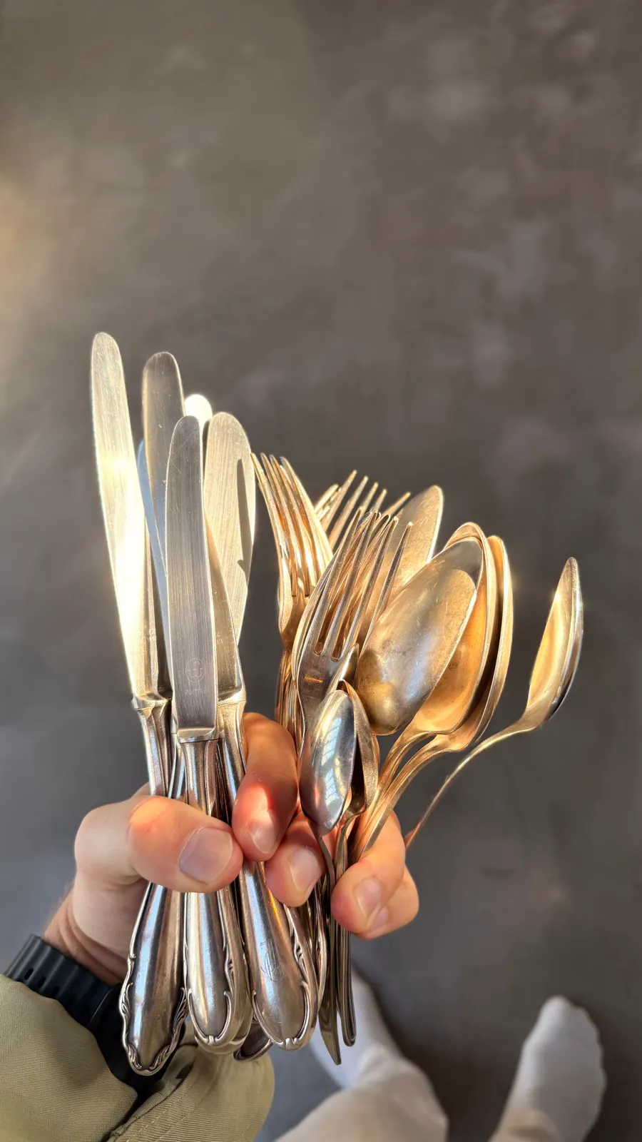 Hand holding a bunch of assorted silver knives, forks, and spoons against a neutral background.