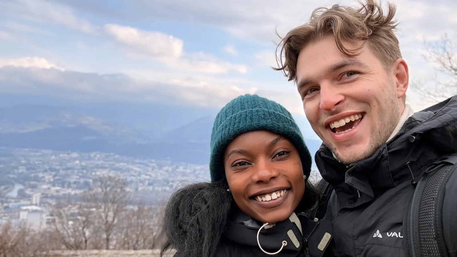 Smiling young couple wearing winter jackets and a beanie, posing for a selfie outdoors with a city and mountains in the background.