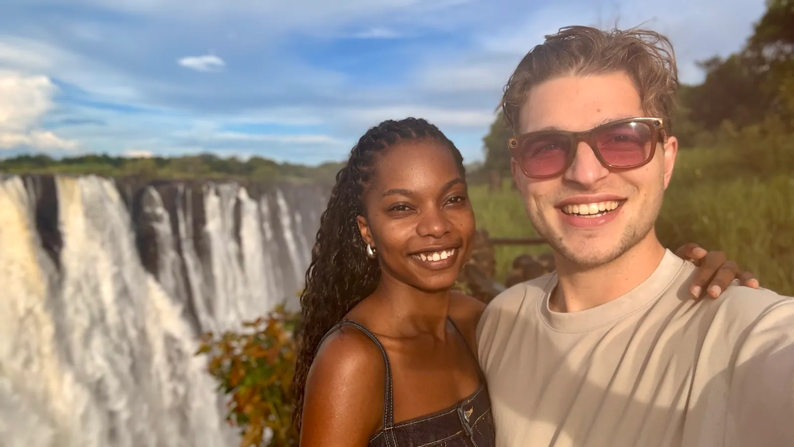 Smiling young couple taking a selfie with a large waterfall and greenery in the background on a sunny day.