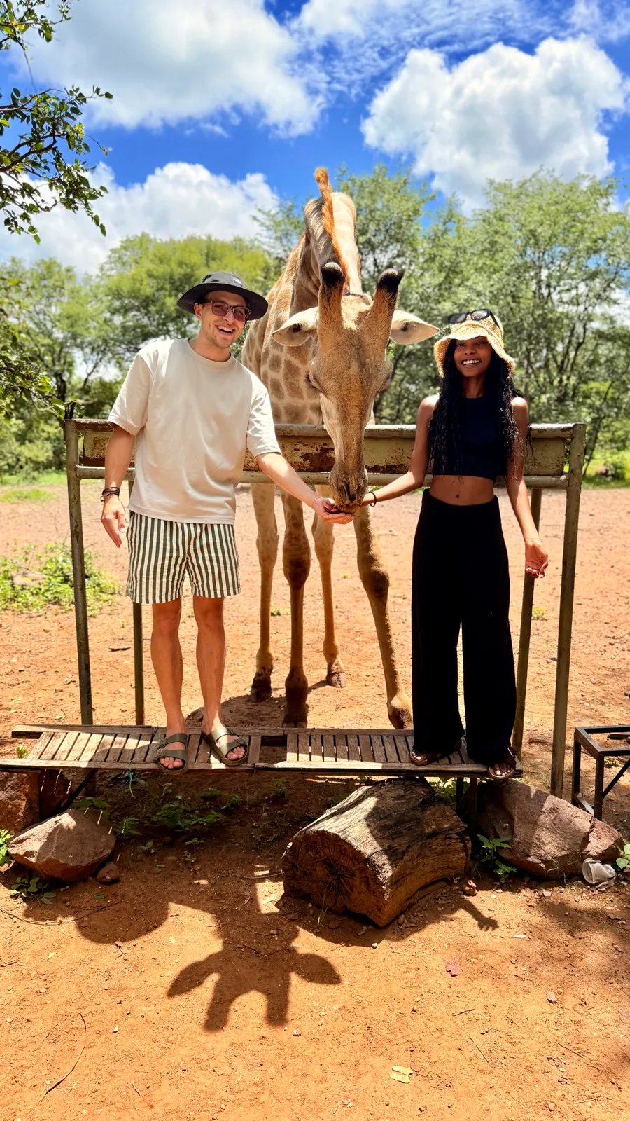 Two people standing on a wooden platform feeding a giraffe that is extending its neck over a metal railing under a blue sky with scattered clouds.