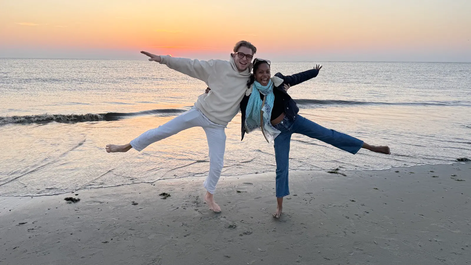 Two people on a beach at sunset, balancing on one leg with arms outstretched, smiling, with the sea and sky in the background.