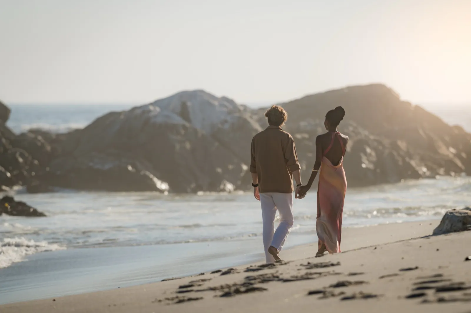 Couple holding hands and walking barefoot on a sandy beach near rocky formations at sunset.