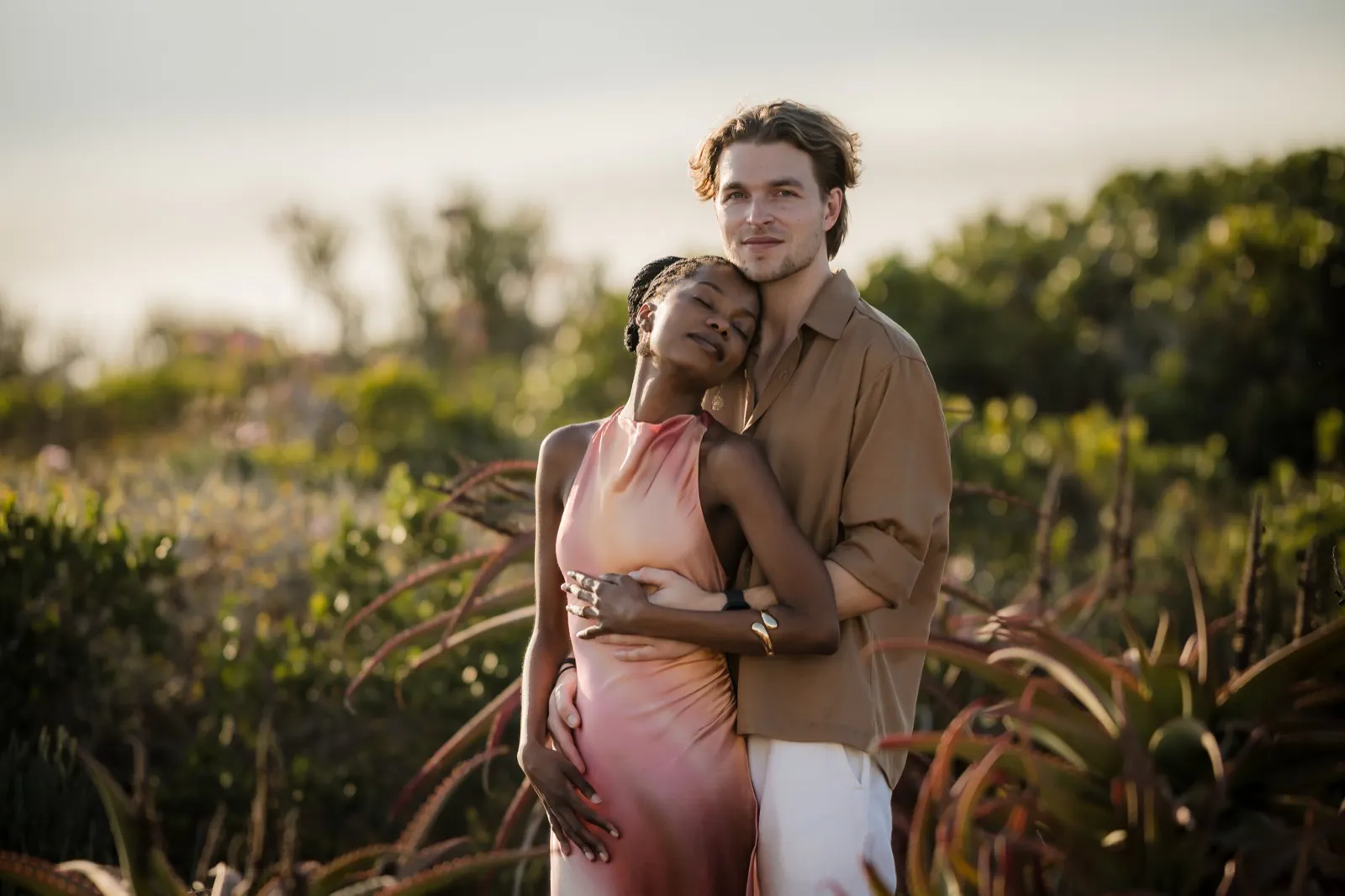Couple standing outdoors with greenery background, woman in sleeveless pink dress leaning her head on man's chest, man embracing her from behind wearing brown shirt and white pants.