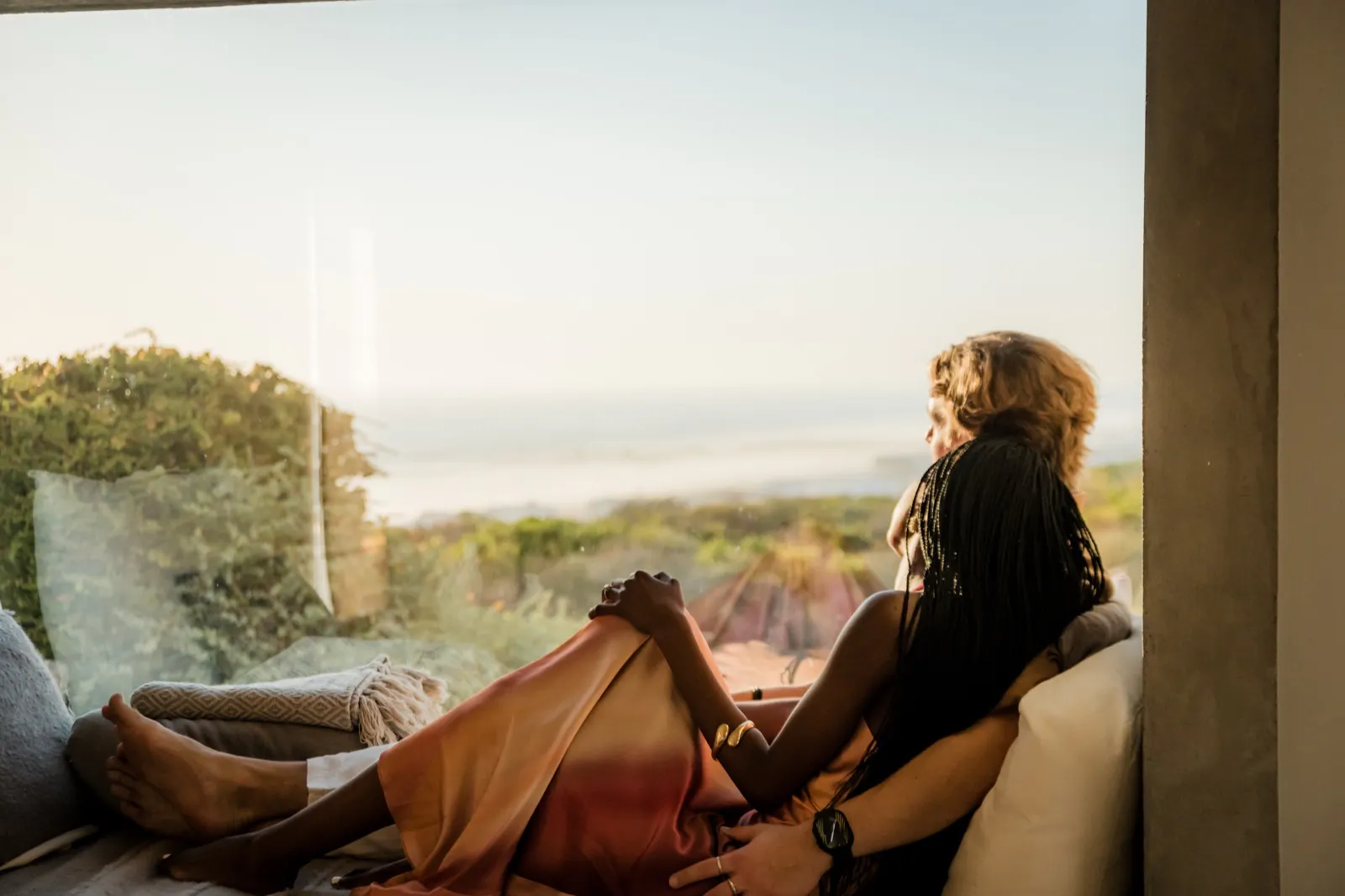 Couple sitting closely together on a couch by a large window, looking out at a scenic, sunlit landscape with greenery and distant water.